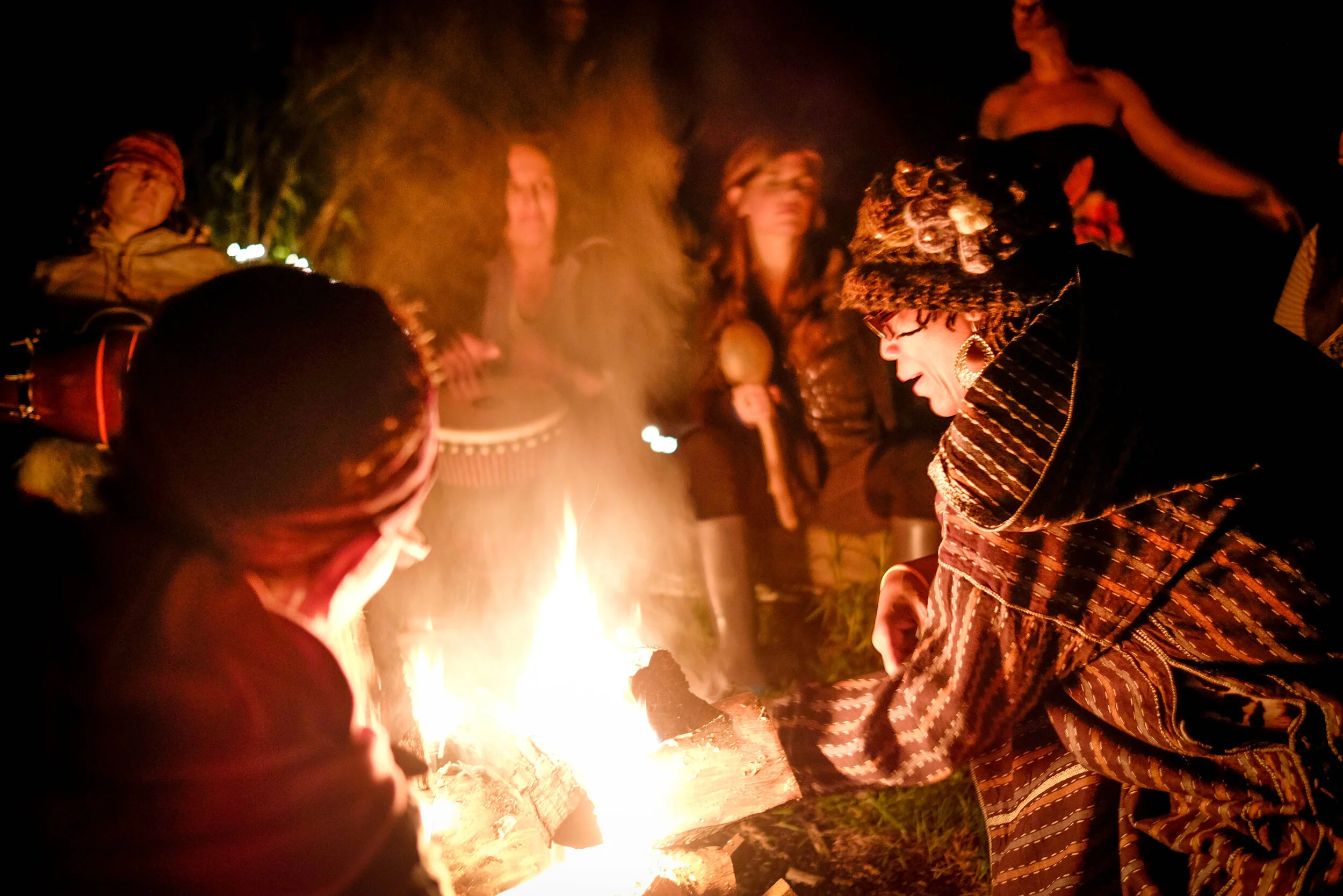 People gathered around a campfire at night, some wearing patterned clothing and headgear, with a woman in a floral hat and glasses leaning toward the fire, and others standing or sitting in the background.