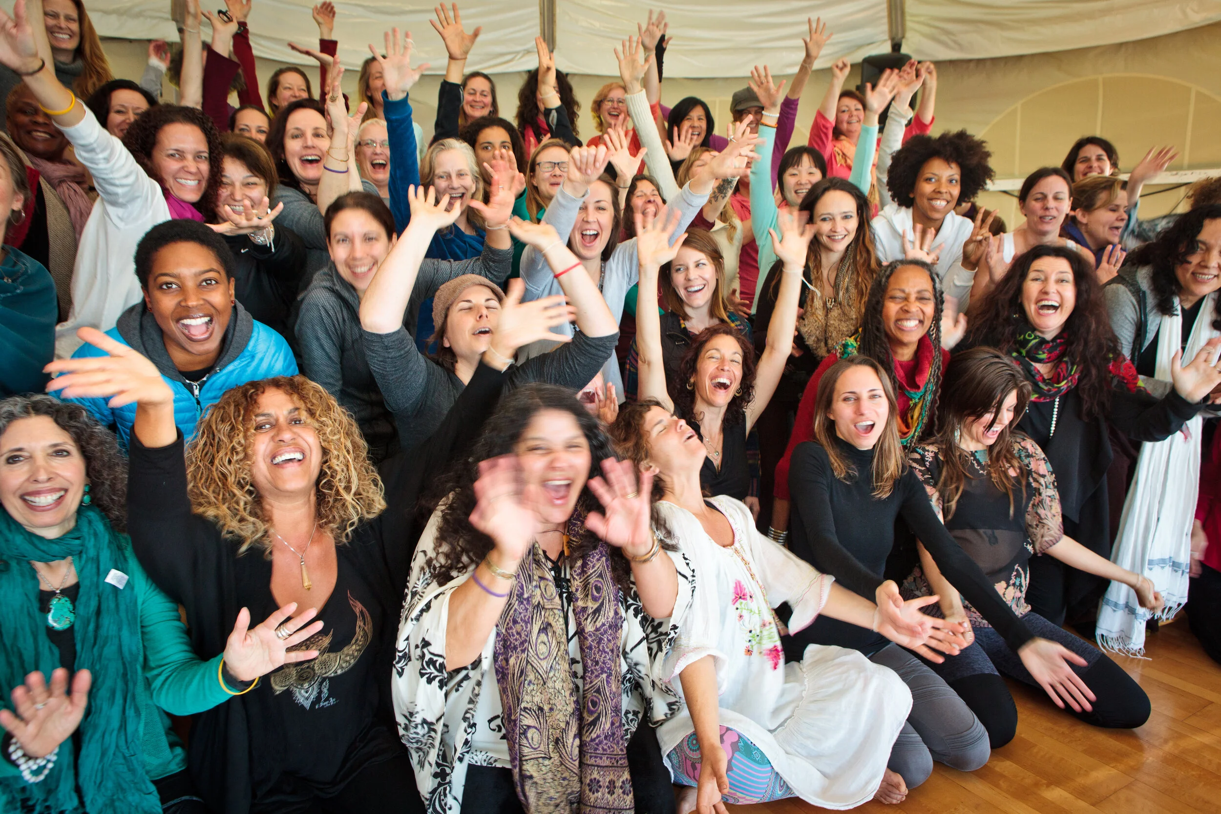 A large group of diverse women smiling and celebrating indoors under a tent at WisdomWomen's Visionary Gathering at Esalen Institute in Big Sur, California.

