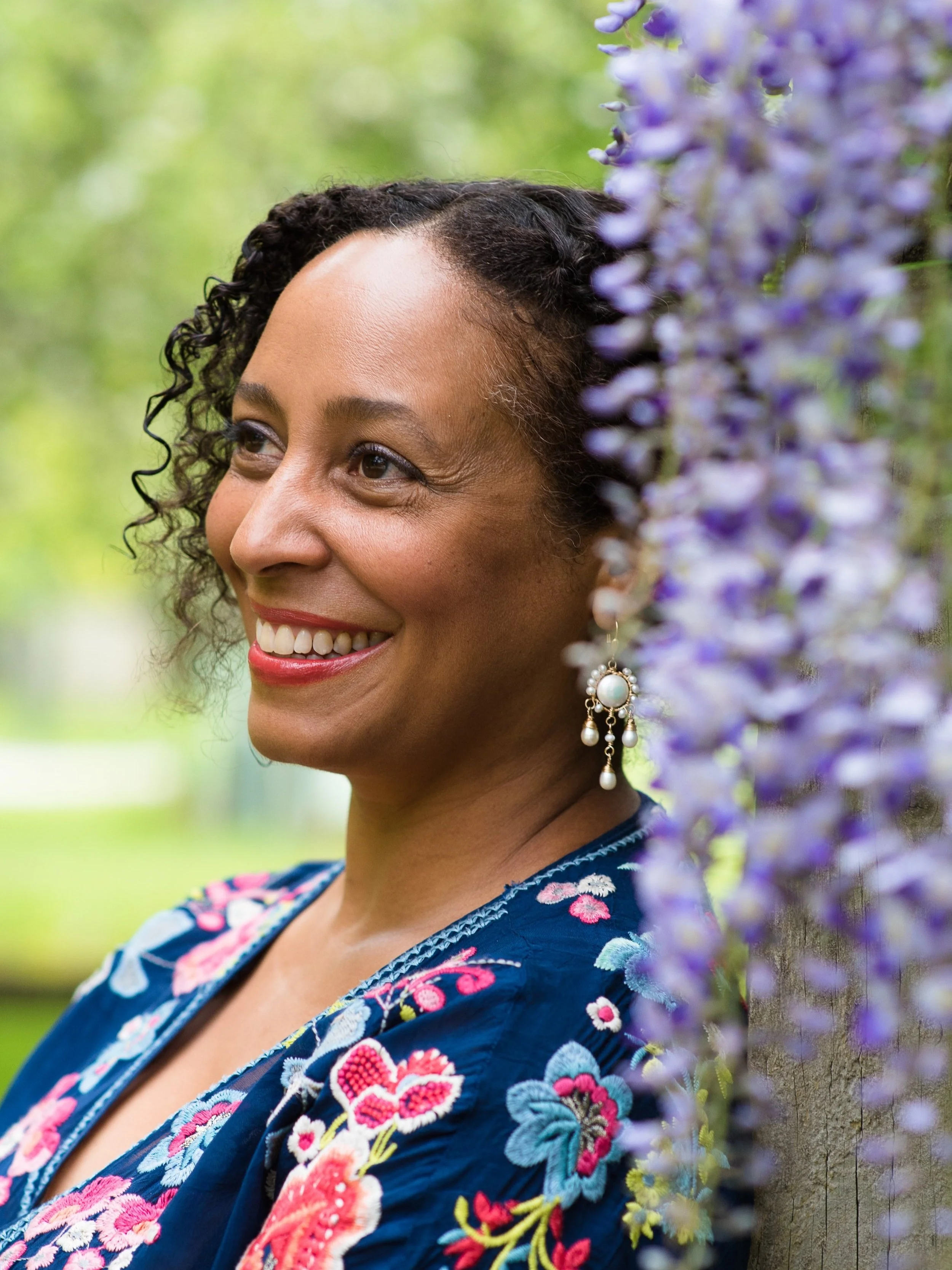 A smiling woman with curly hair is standing near a wooden post decorated with purple flowers. She is wearing a blue embroidered top and pearl earrings, with a blurry green outdoor background.