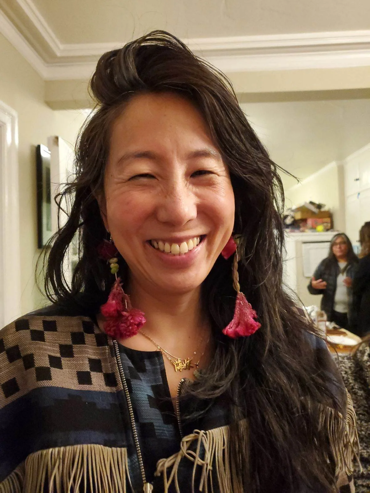 A woman with dark, wavy hair smiling at the camera, wearing large pink earrings and a patterned top, in an indoor setting with other people in the background.