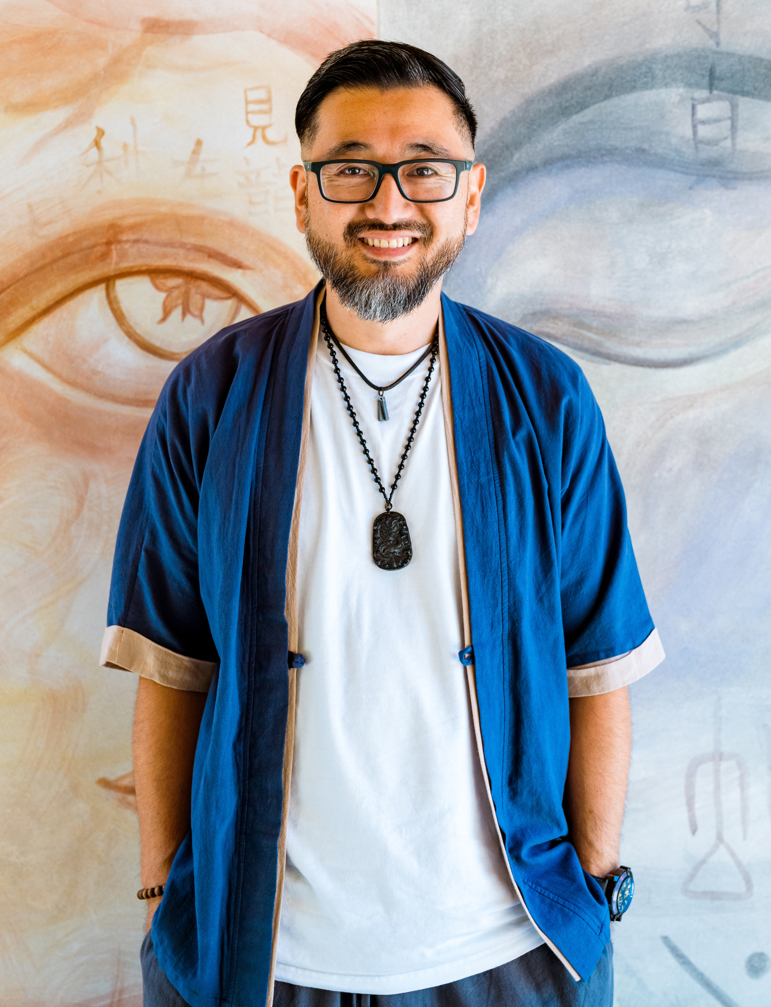 A smiling man with glasses, a beard, and short black hair, wearing a white t-shirt and blue open shirt with rolled-up sleeves, standing in front of a painted mural with eyes and symbols.