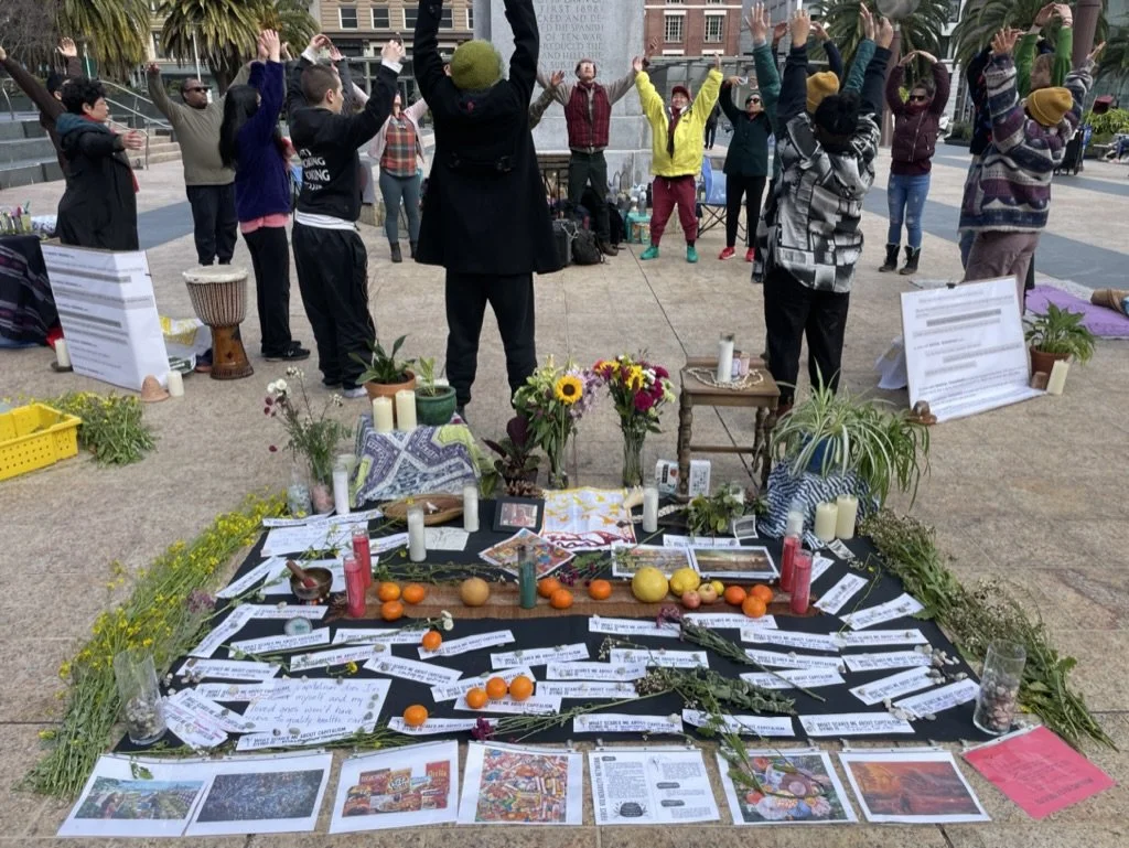 A group of people in a city park raising their hands in a circle, surrounded by an altar with flowers, candles, oranges, papers, and photographs.