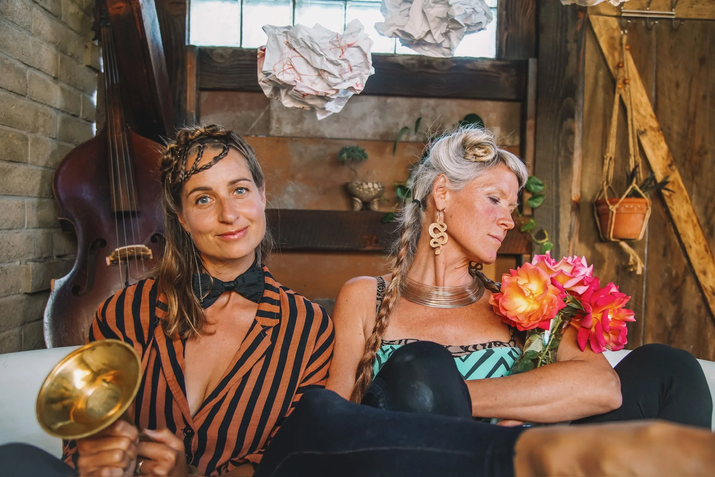 MaMuse on white couch in a rustic room with brick and wooden walls. The woman on the left wears a striped shirt with a bow tie, smiling at the camera. The woman on the right, with braided gray hair and large earrings, is holding a bouquet of flowers.