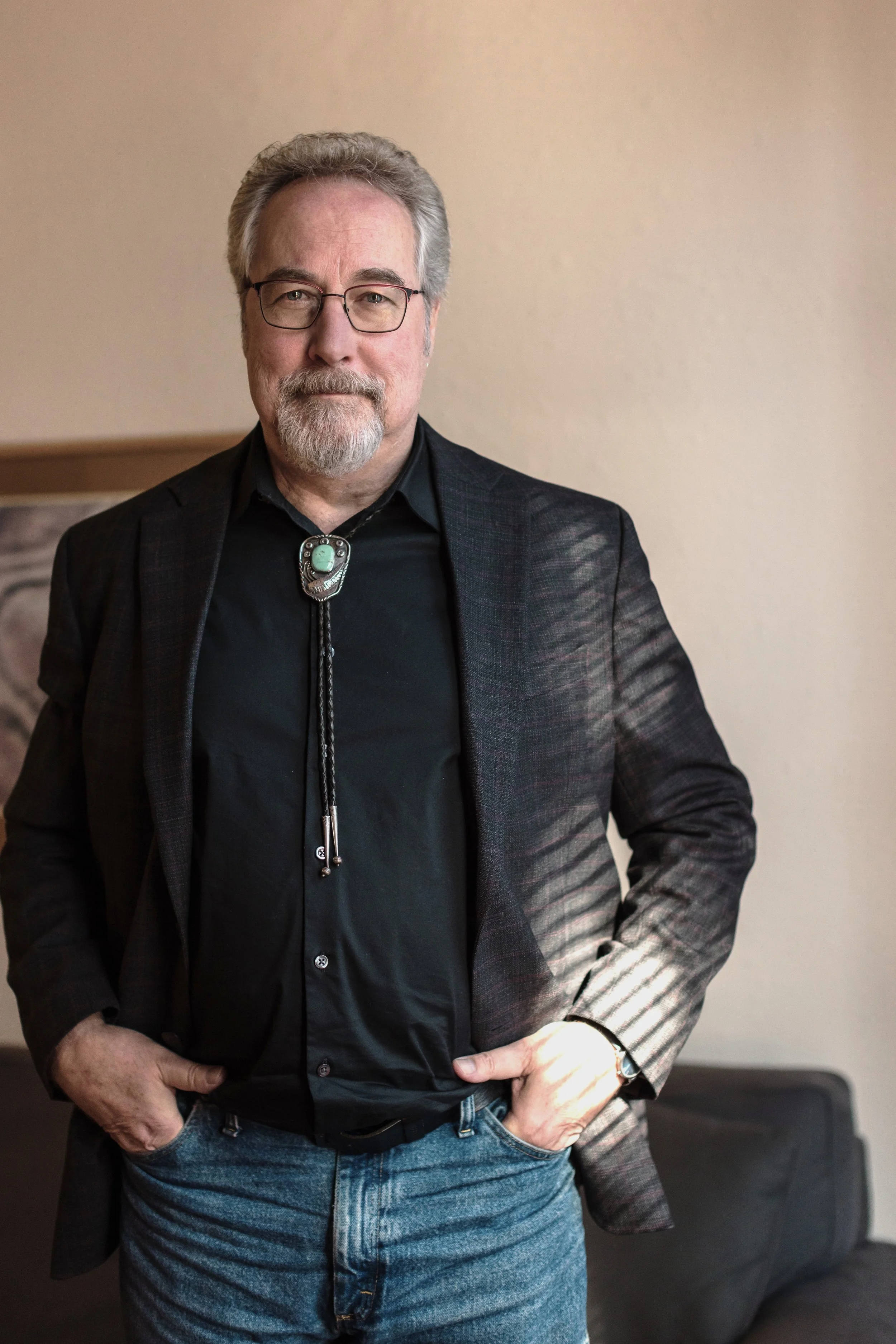 A photo of Francis Weller wearing glasses wearing a black shirt, blazer, and jeans, standing with hands in pockets, in a room with neutral-colored walls.