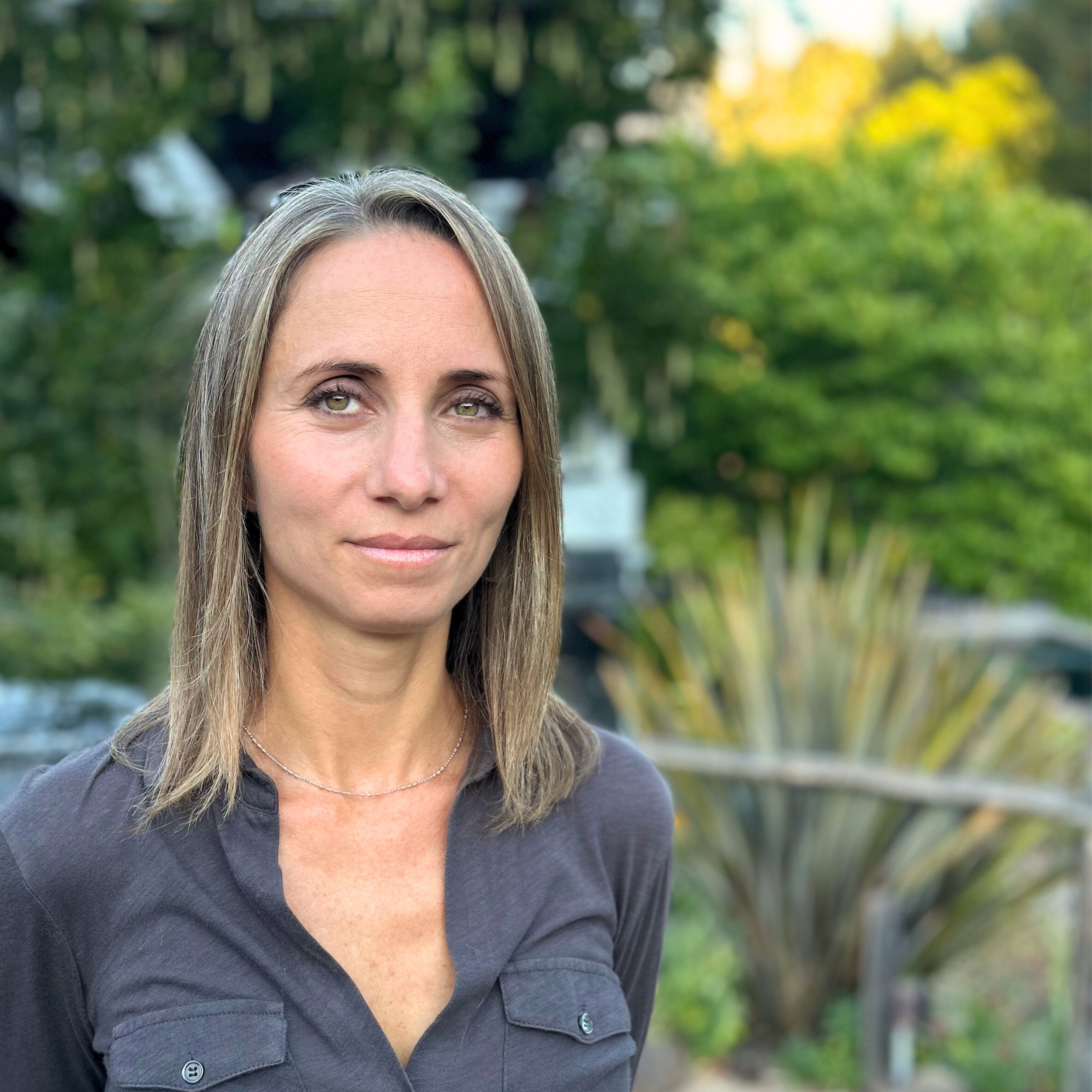 A photo of Michelle Stransky who has light brown, shoulder-length hair and green eyes standing outdoors with trees and plants in the background.