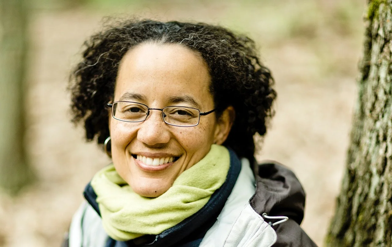 A photo of Kaira Jewel Lingo smiling  with curly hair, glasses, and a green scarf outdoors in a wooded area.