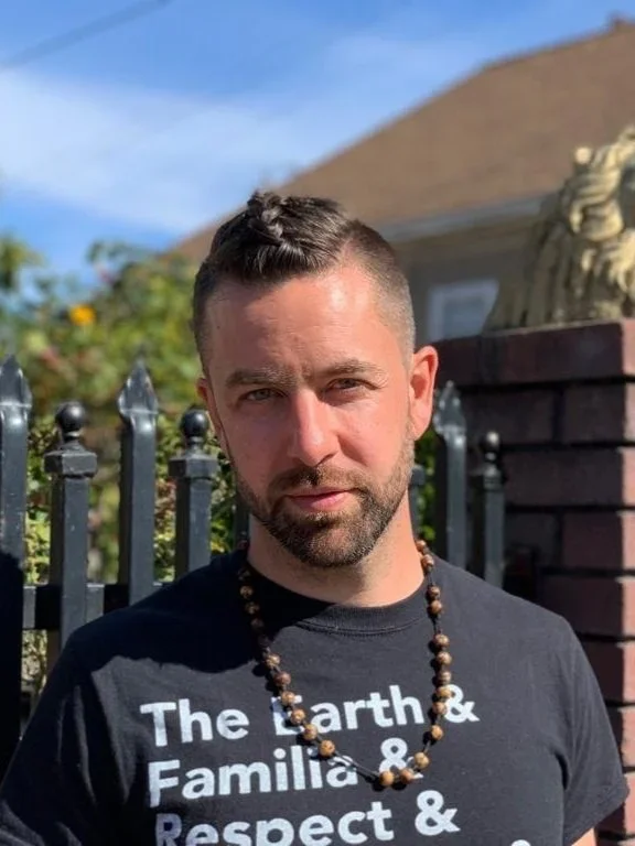 Lu Aya with short dark hair and a beard, wearing a black t-shirt with white text and a beaded necklace, standing outdoors in front of a black fence and brick wall, with trees and a house in the background.