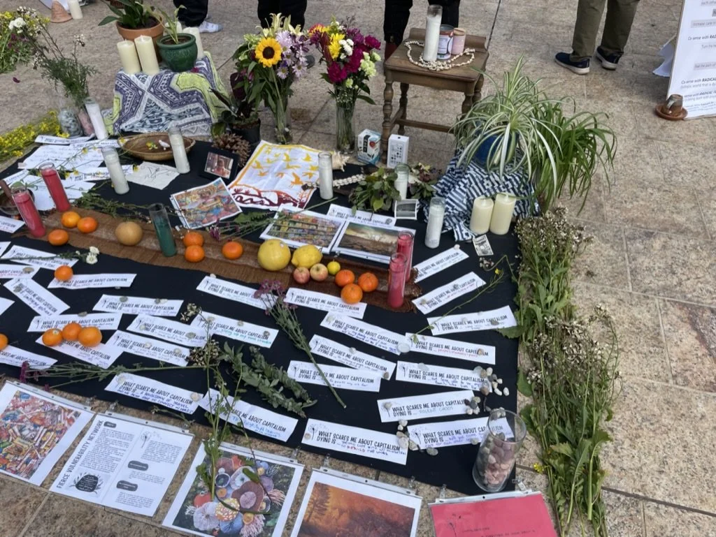 A memorial or ofrenda setup with flowers, candles, photographs, oranges, apples, and papers on a black cloth, surrounded by plants and candles.