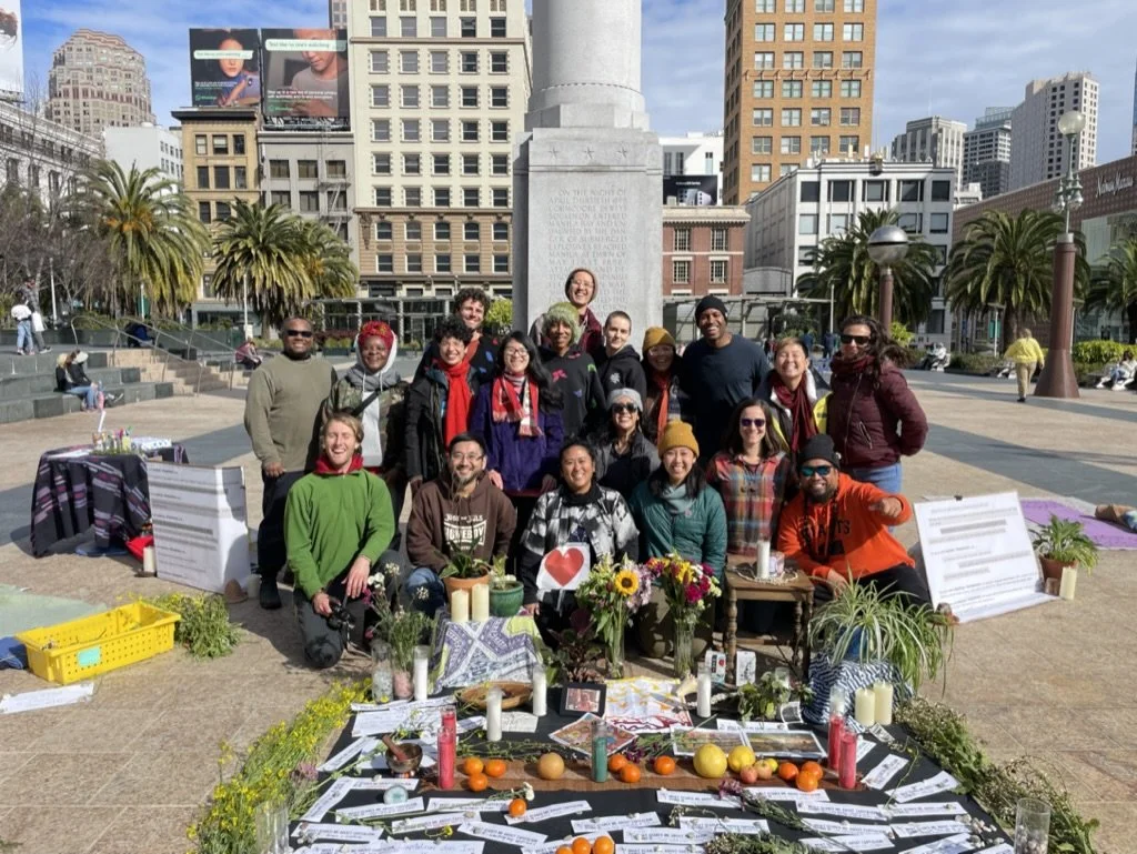 A diverse group of people gathered around a display of flowers, candles, and produce in an urban park with skyscrapers and palm trees in the background.