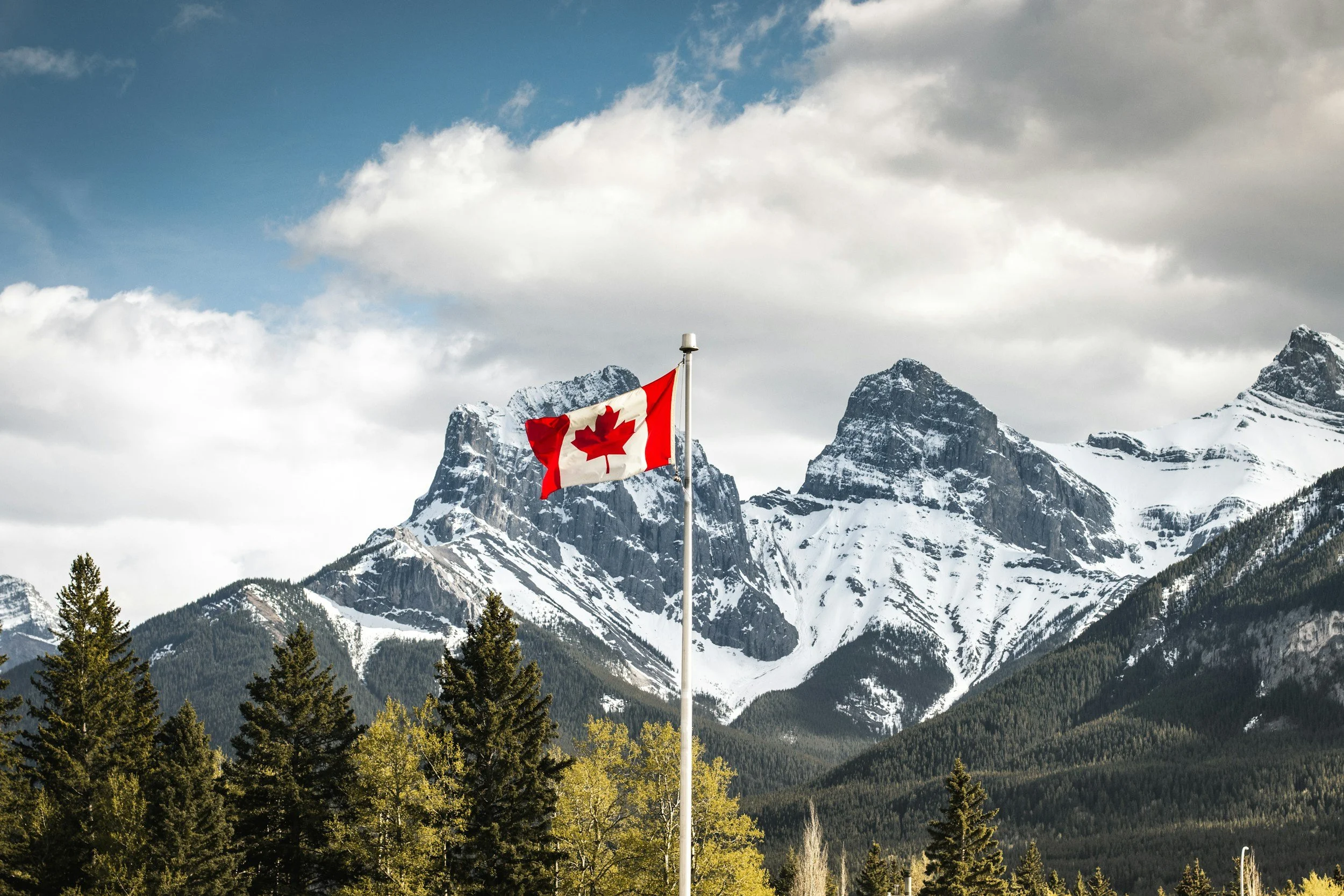 Canadian flag flying in front of snow-capped mountains and green pine trees under a cloudy sky.