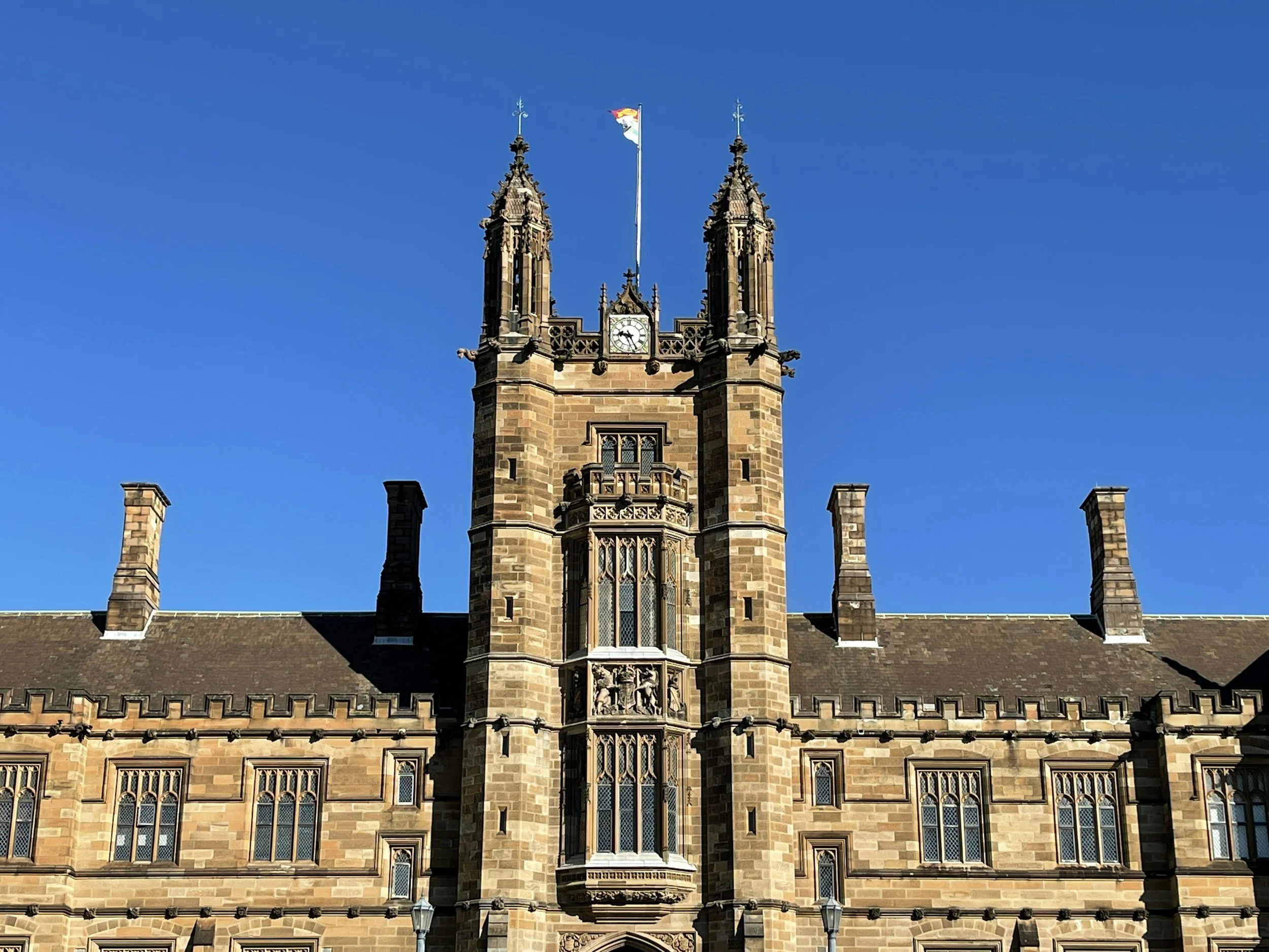 Sydney University building with a central tower featuring a clock, two spires, and stained glass windows, set against a clear blue sky.