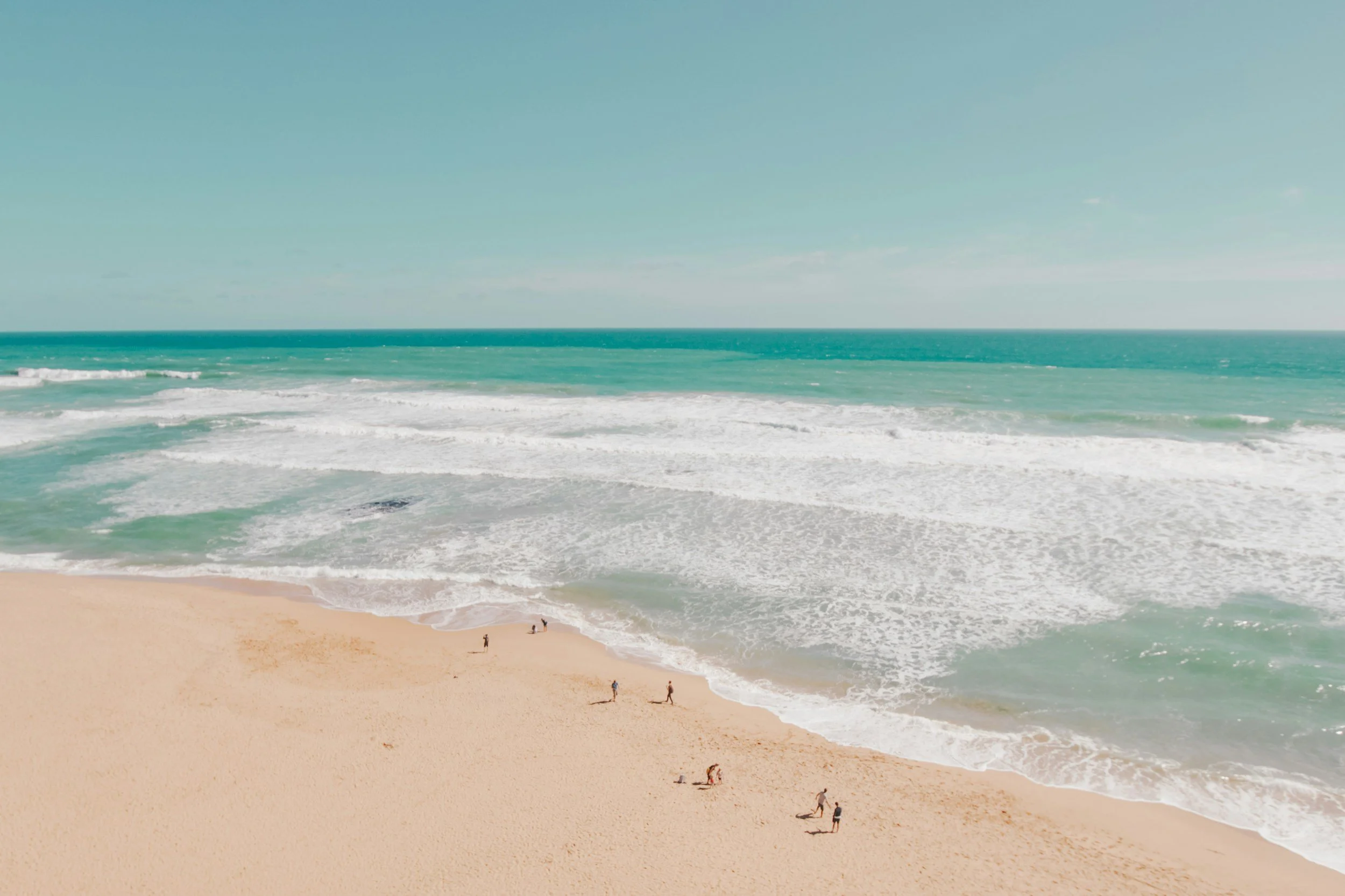 Aerial view of a sandy beach in Australia with a few people walking along the shoreline. The ocean waves are rolling in with turquoise water under a clear sky.