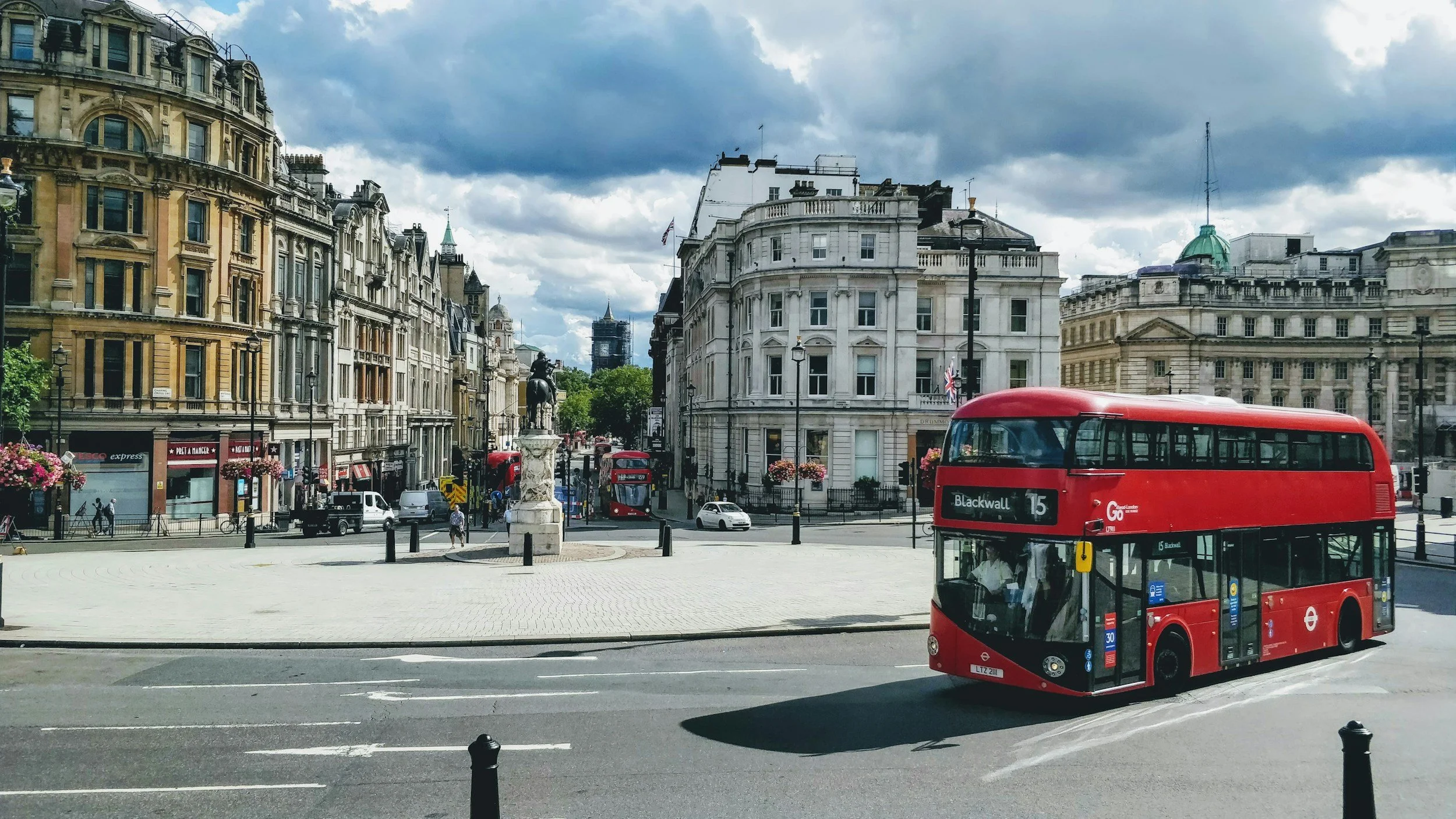 London UK double-decker bus driving on a city street with historic buildings and a statue in the background, and cloudy skies overhead.