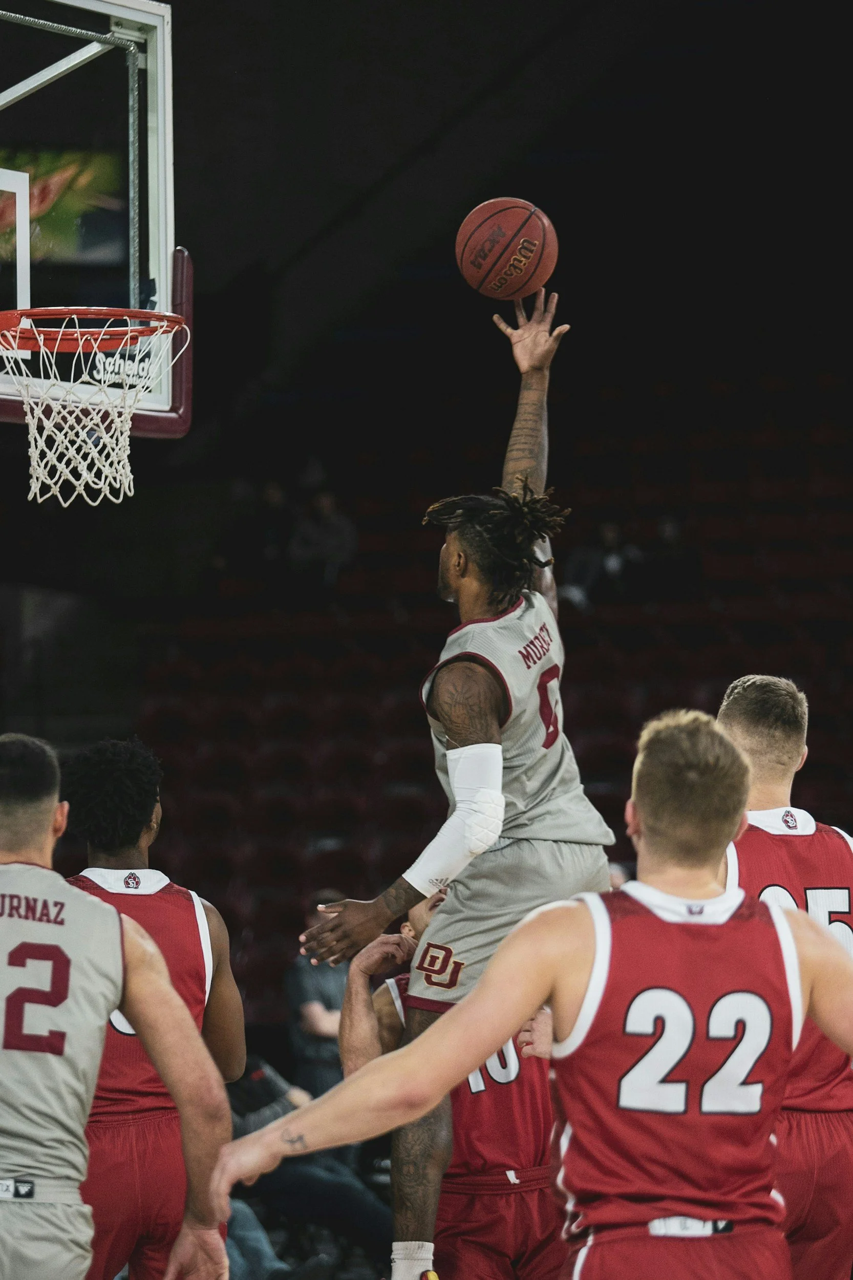 A American college basketball player in a white and red uniform jumping to shoot the ball towards the hoop, while other college players observe.