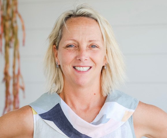 A smiling woman with blonde hair, wearing a sleeveless top, standing indoors near a light-colored wall.