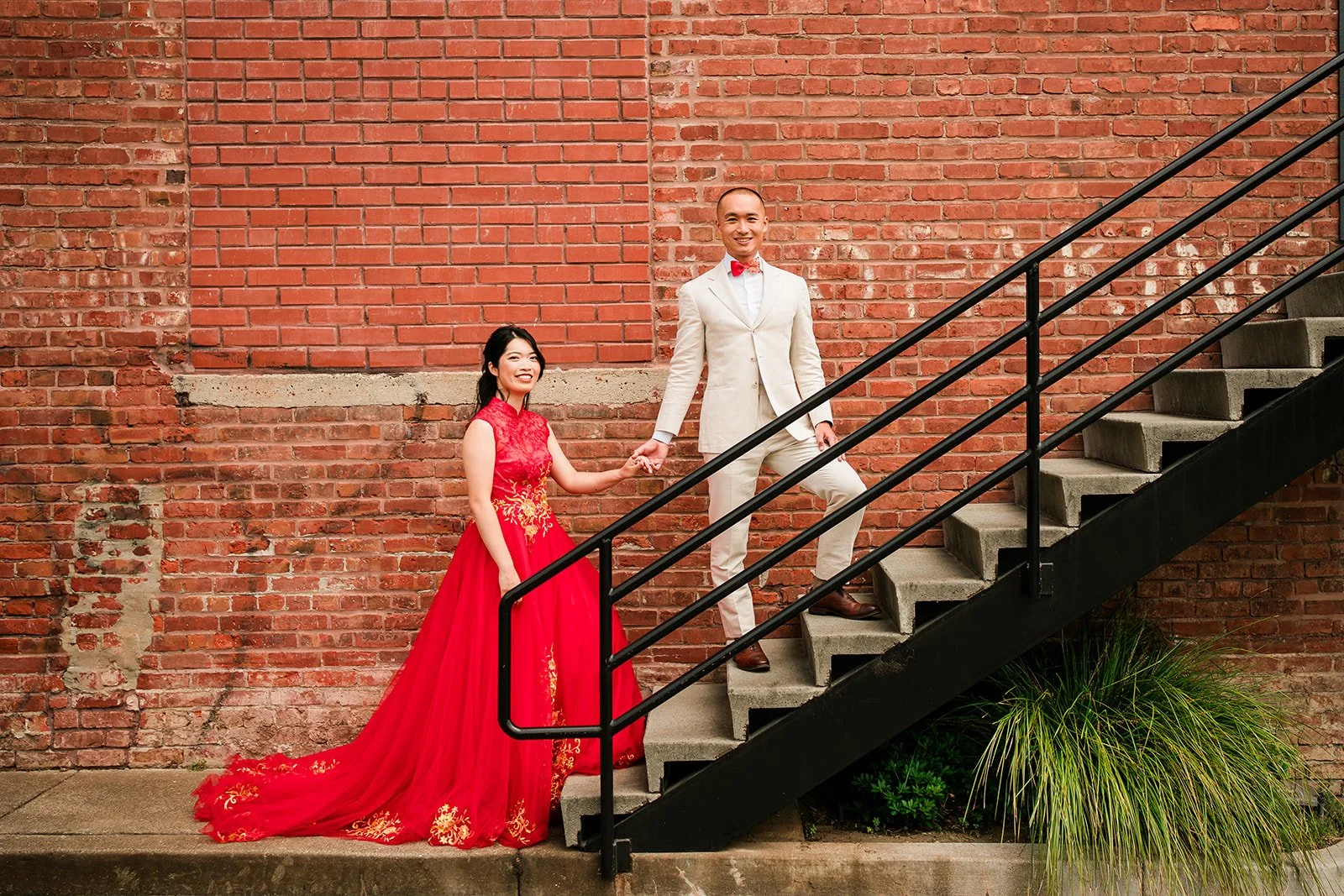 A couple holding hands on a staircase against a red brick wall, with the woman in a red dress and the man in a beige suit and red bow tie.