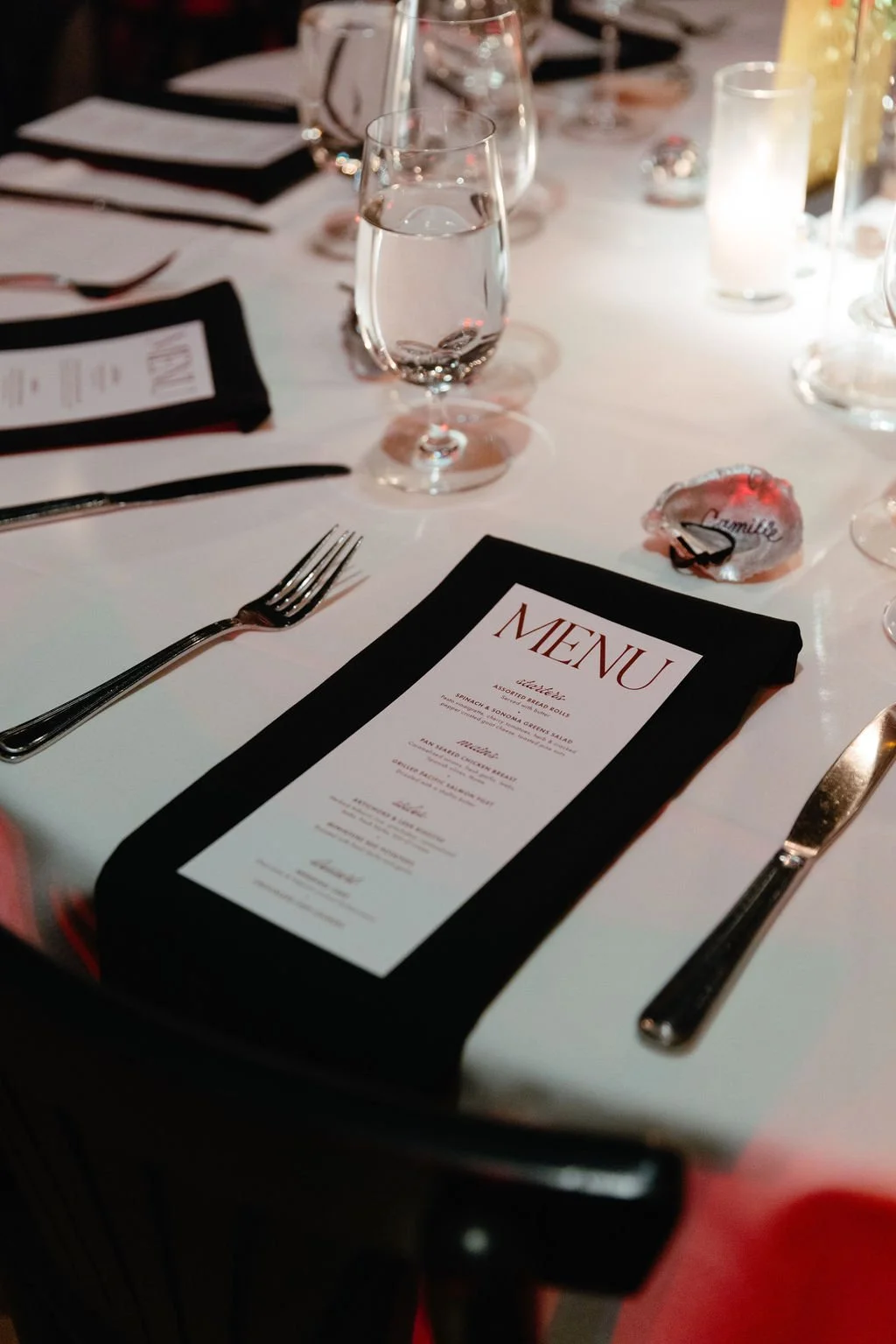 A table set for a formal event with a black-bordered menu, a fork on the left, and a knife on the right. There are water glasses, candles, and a small decorative name tag on the table.