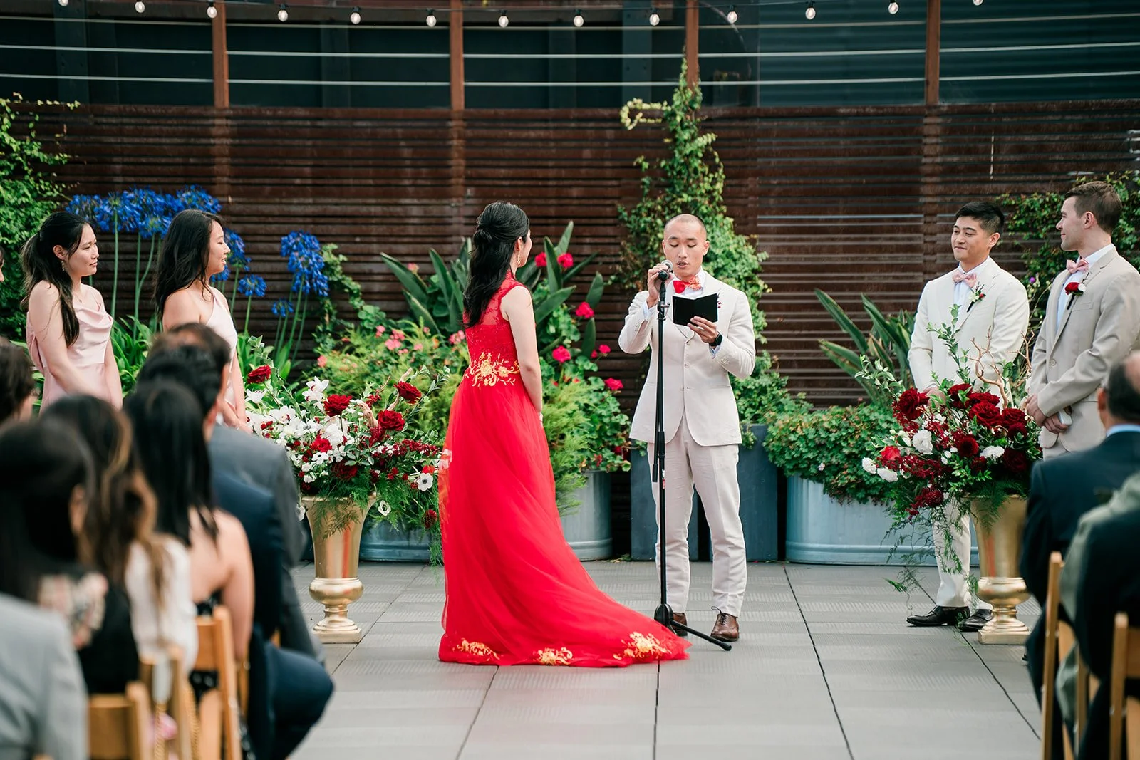 A wedding ceremony taking place outdoors on a decorated patio with floral arrangements, where a woman in a red dress is speaking into a microphone, while a man in a beige suit and another man in a white suit listen. Bridesmaids and groomsmen, dressed