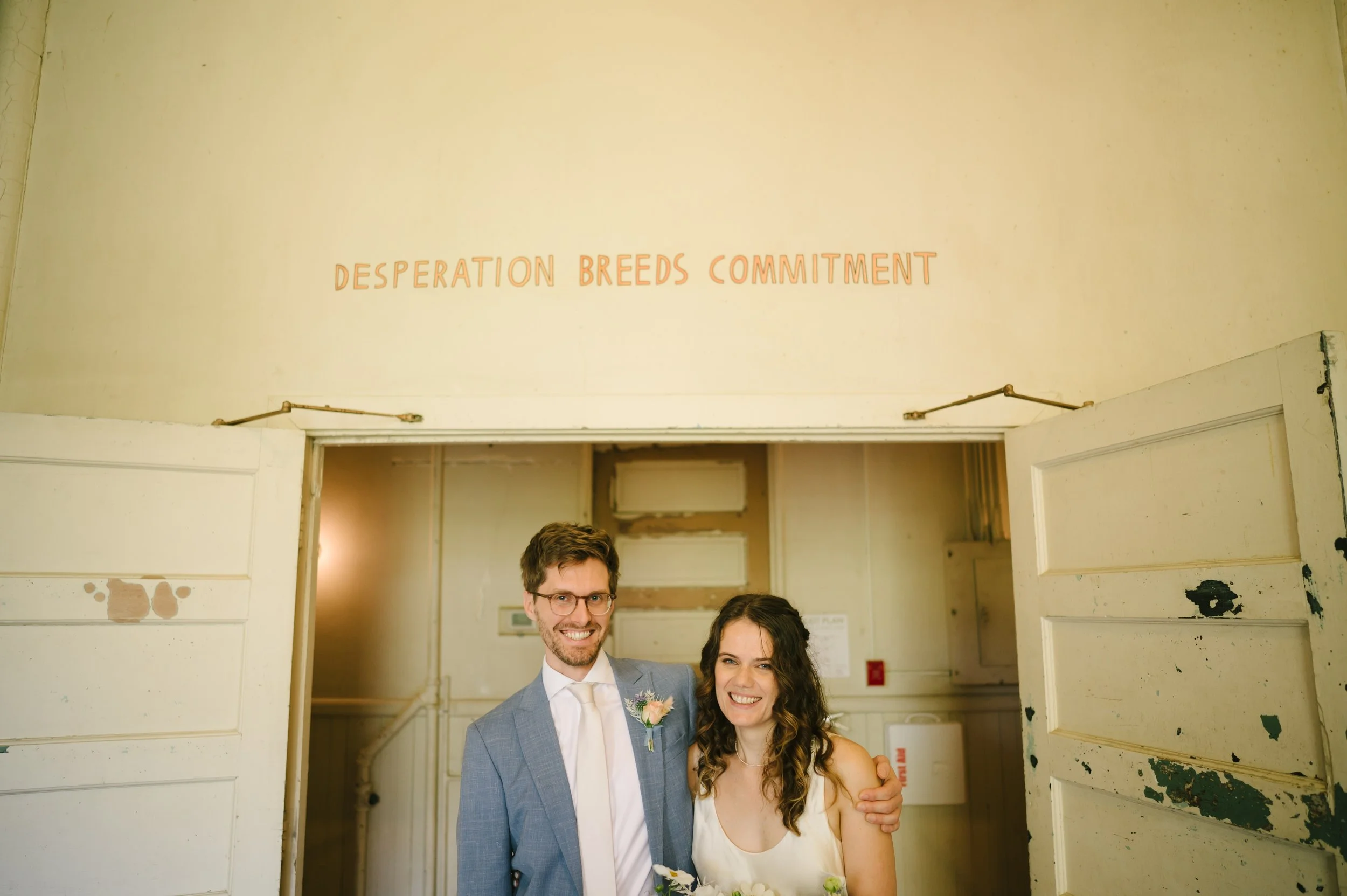 A smiling couple dressed in wedding attire standing in front of a doorway, with the man wearing a light gray suit and the woman in a white dress, holding a bouquet of flowers.