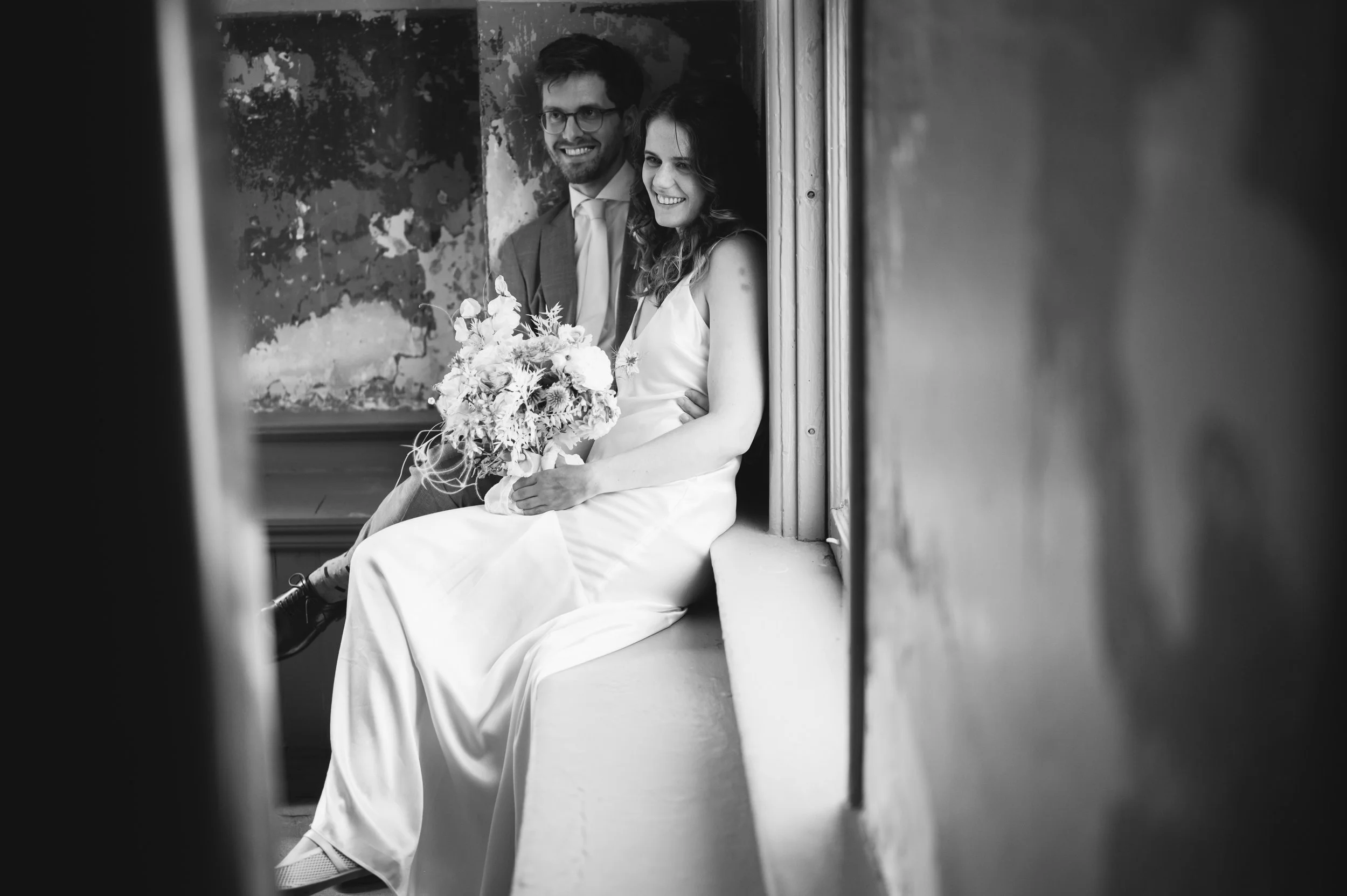 A smiling bride in a white dress holding a bouquet, sitting on a windowsill beside a groom in a suit. Both are looking out a window with an old wall in the background.
