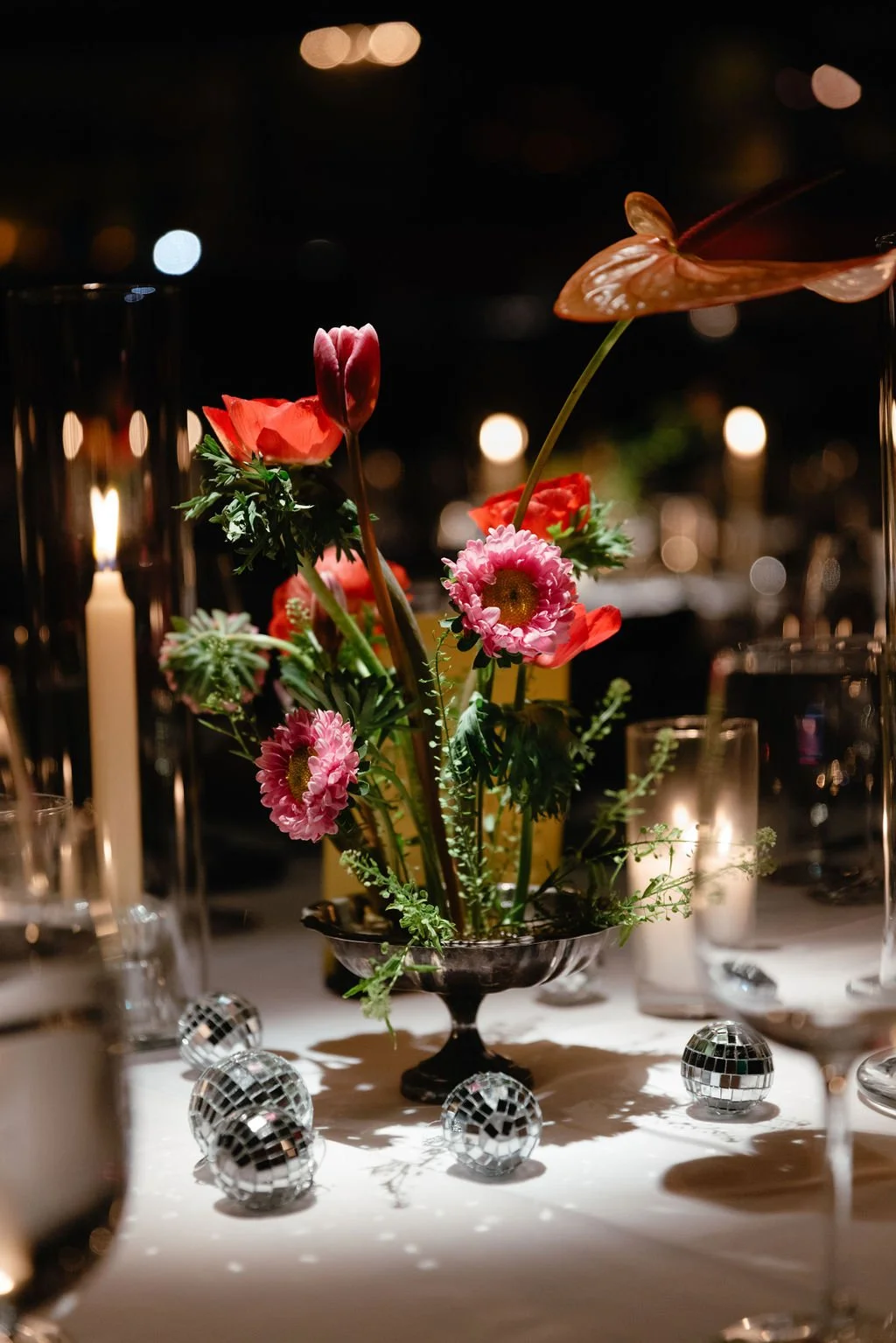 A floral centerpiece with pink gerbera daisies, red poppies, and anthurium in a silver bowl, surrounded by candles and mini disco balls on a table at night.