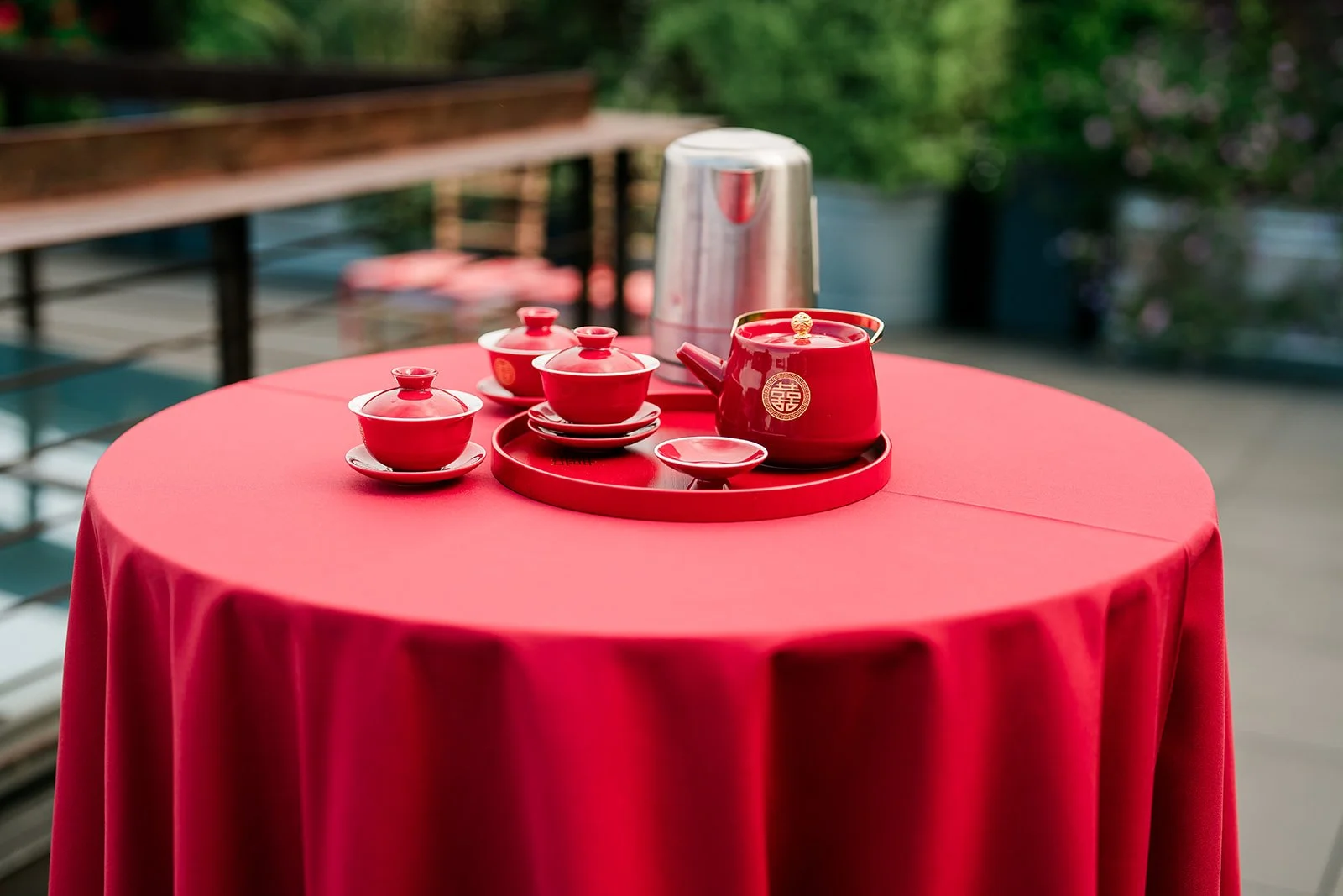 Red teapot and matching cups with saucers on a round red tablecloth, outdoor setting with blurred greenery and wooden railing in background.