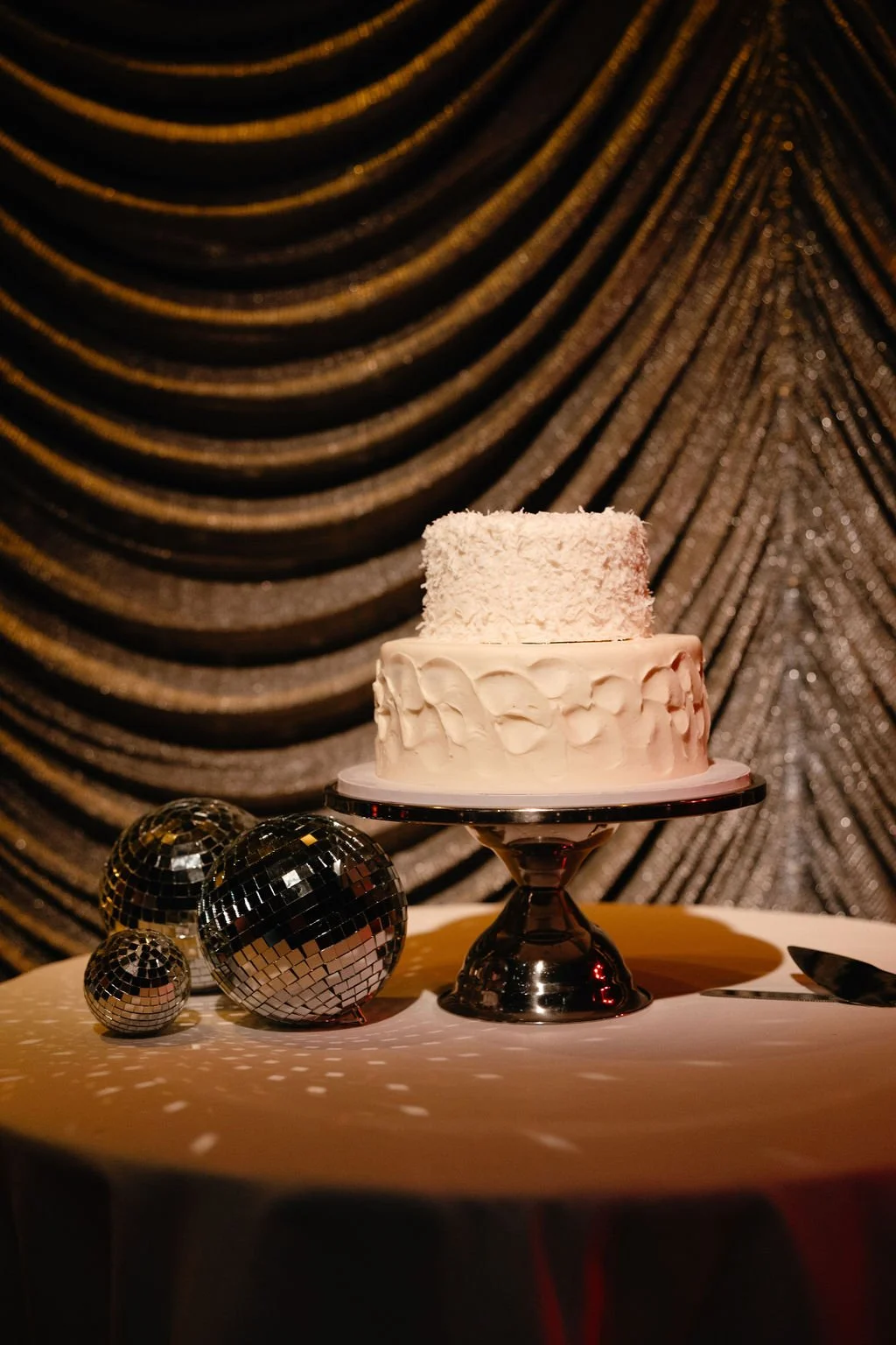A cake on a silver cake stand decorated with white frosting and coconut on top, with three disco balls of different sizes on the table, set against a dark, glittery, gold and black draped backdrop.