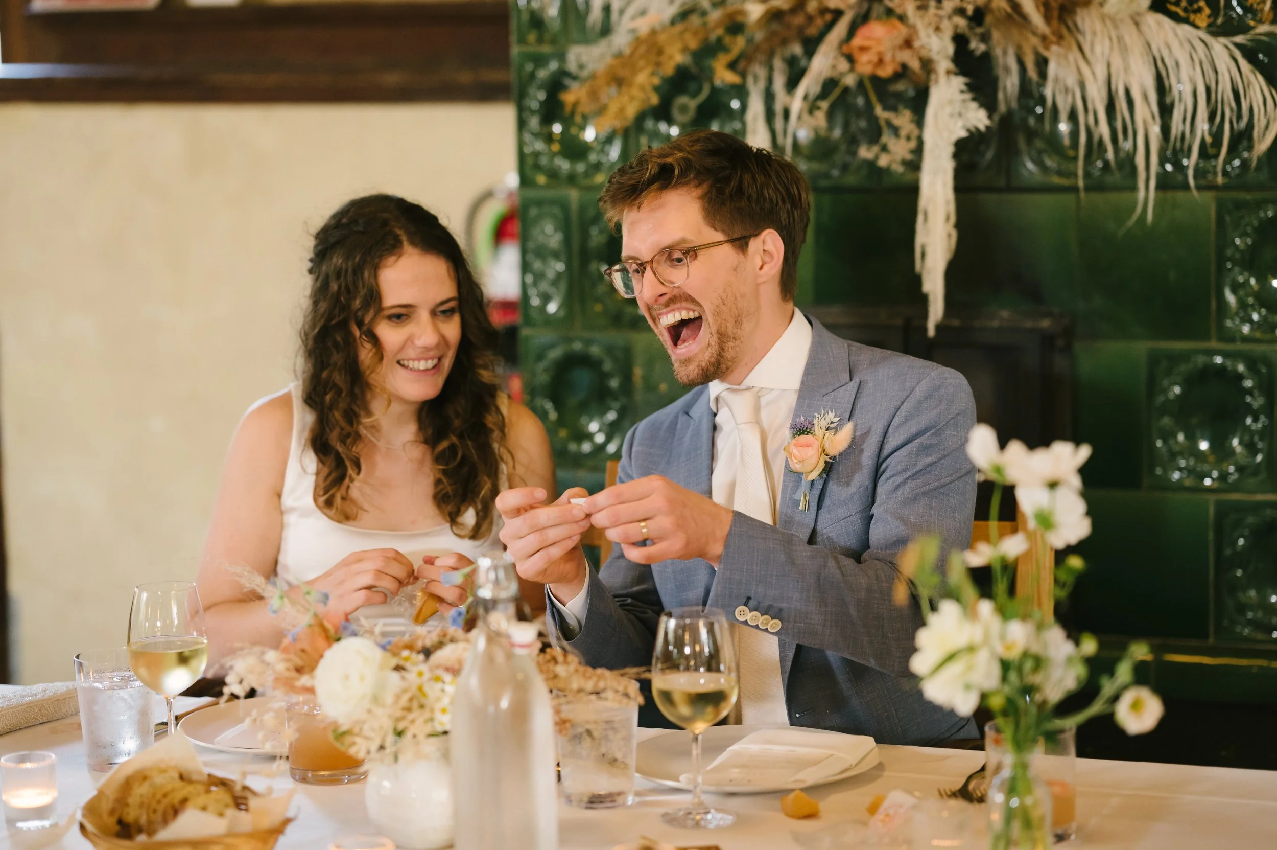 A groom and bride smiling and opening wedding gifts at a sweetheart table decorated with flowers, wine glasses, and tableware, at the Marin Headlands Center for the Arts.