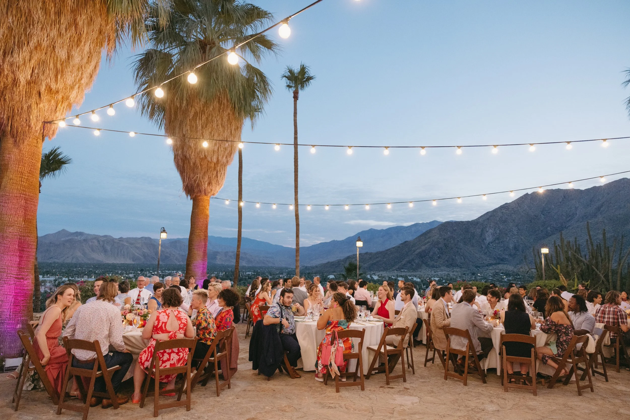Outdoor evening social gathering with round tables, surrounded by people, under string lights, with large palm trees and mountain landscape in the background.