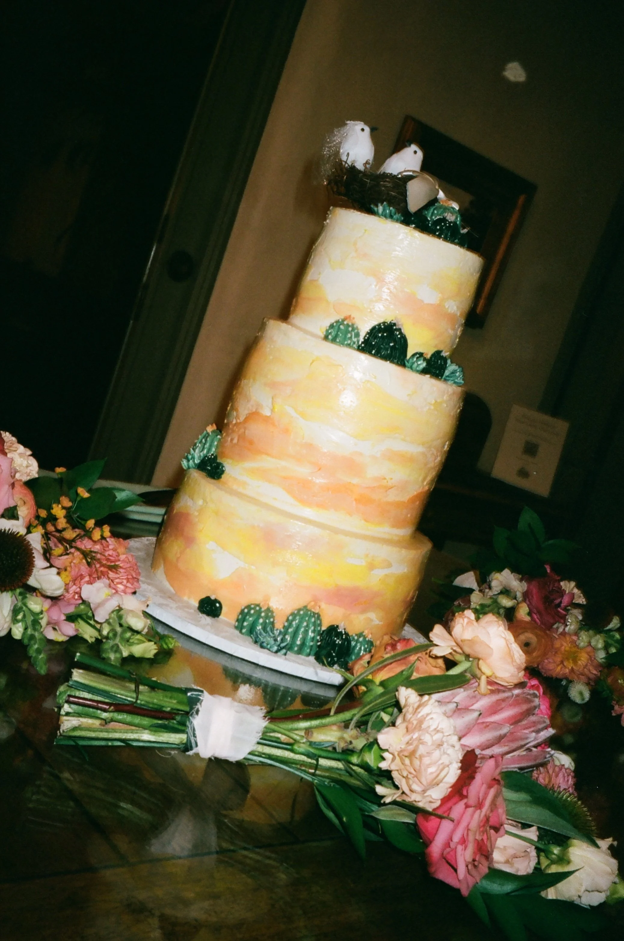 Three-tiered wedding cake with a desert sunrise watercolor design, decorated with small green cacti and topped with a rustic nest with eggs and two small birds, surrounded by pink and white flowers.