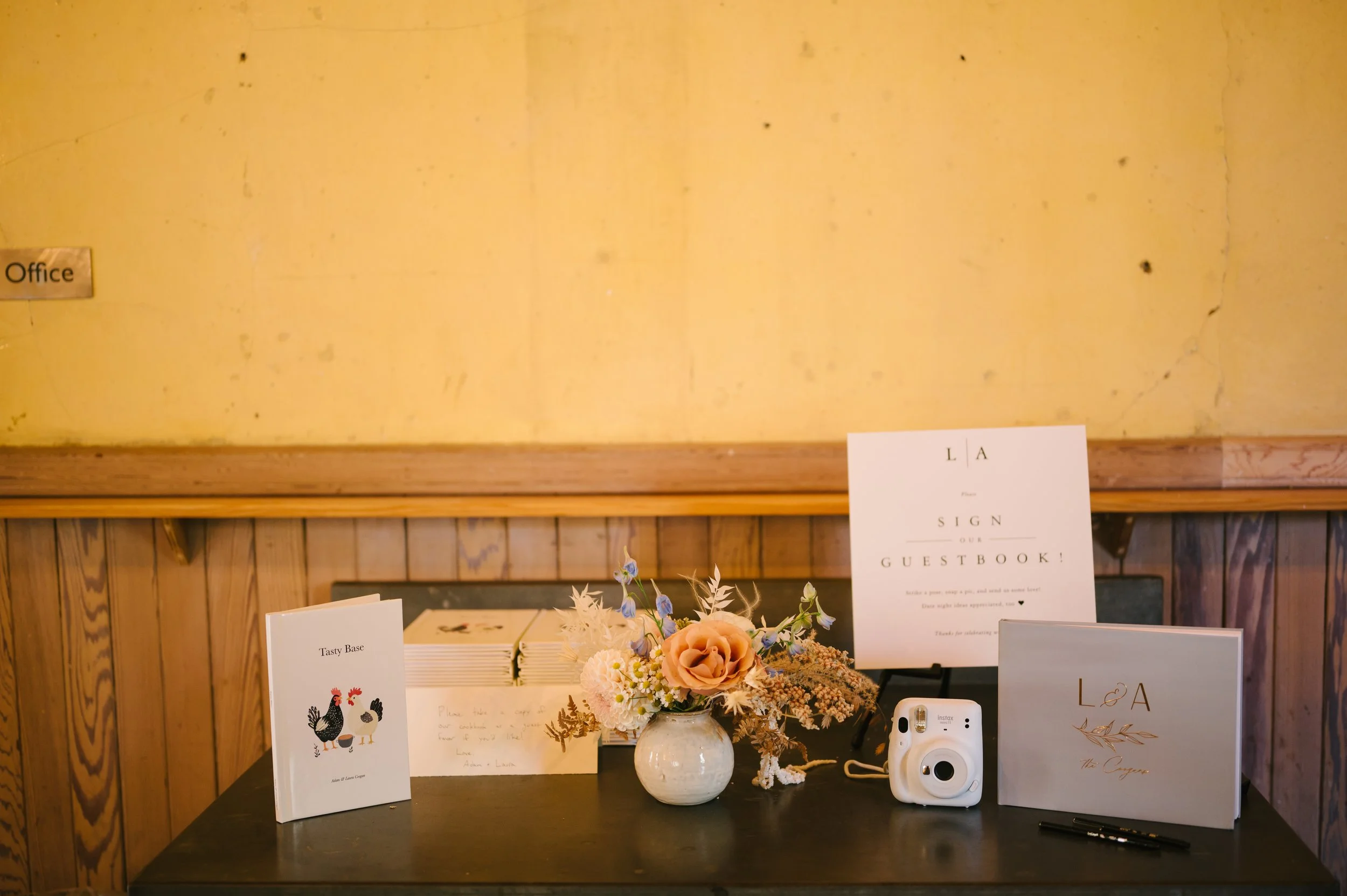 Table with guestbook, flowers, camera, pens, and signs, against yellow wall with wood paneling, in a cozy indoor setting.