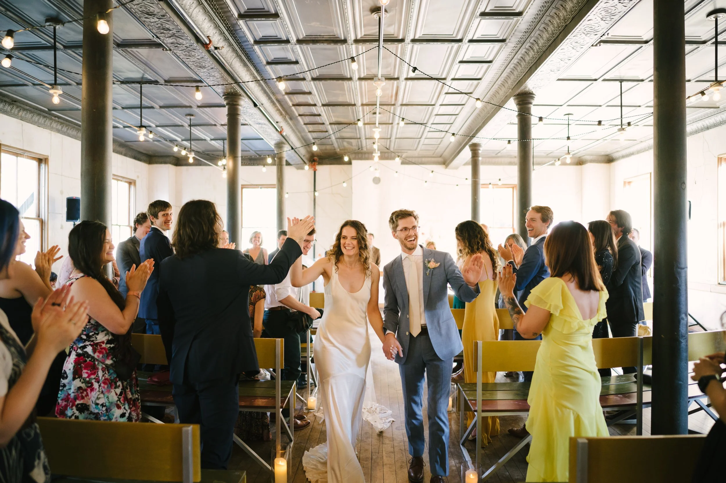 A newlywed couple walking hand in hand down the aisle, smiling, surrounded by guests clapping and cheering inside a spacious, well-lit wedding venue with high ceilings and string lights.