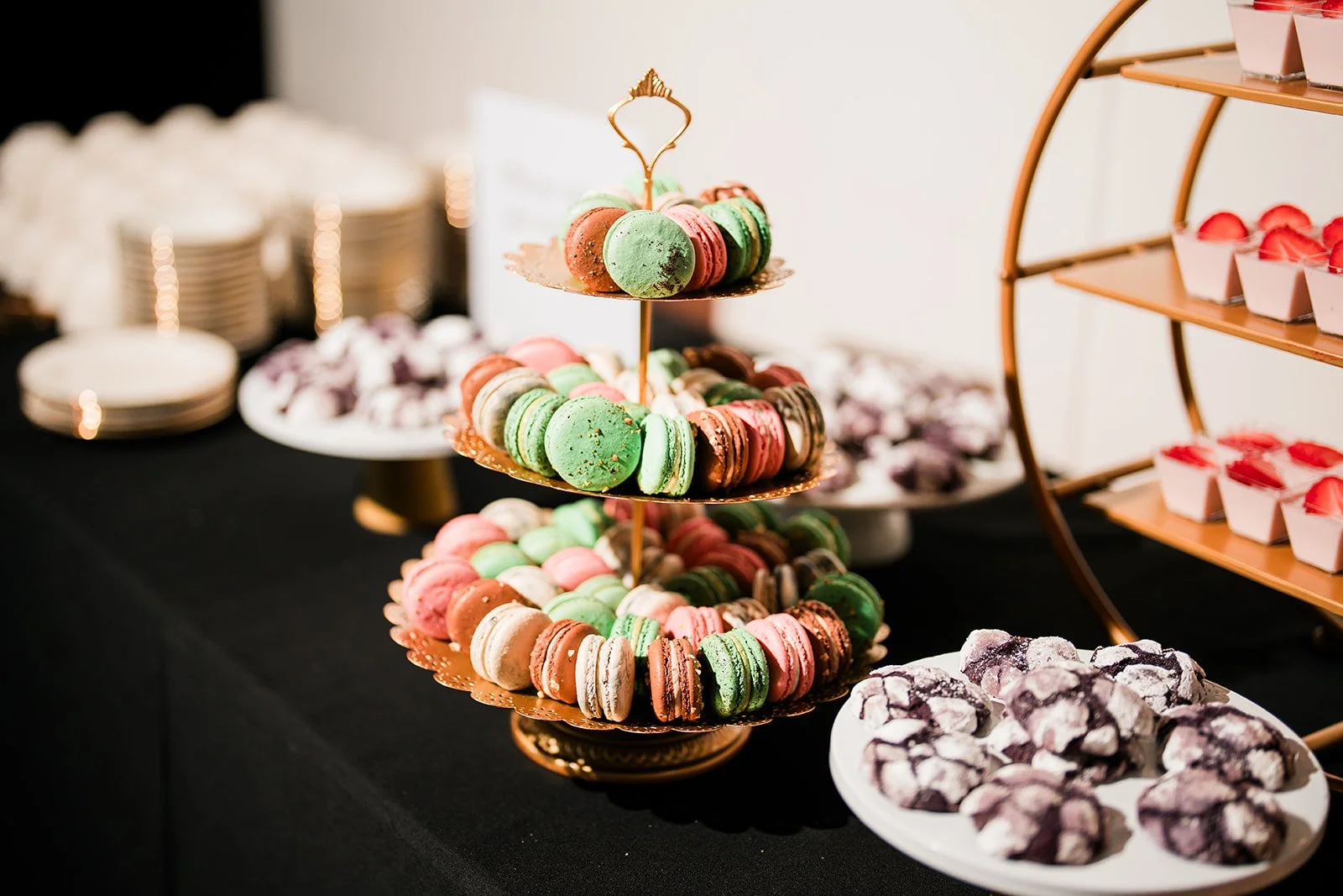 Assorted colorful macarons on a multi-tiered stand, with plates of cookies and other desserts in the background.