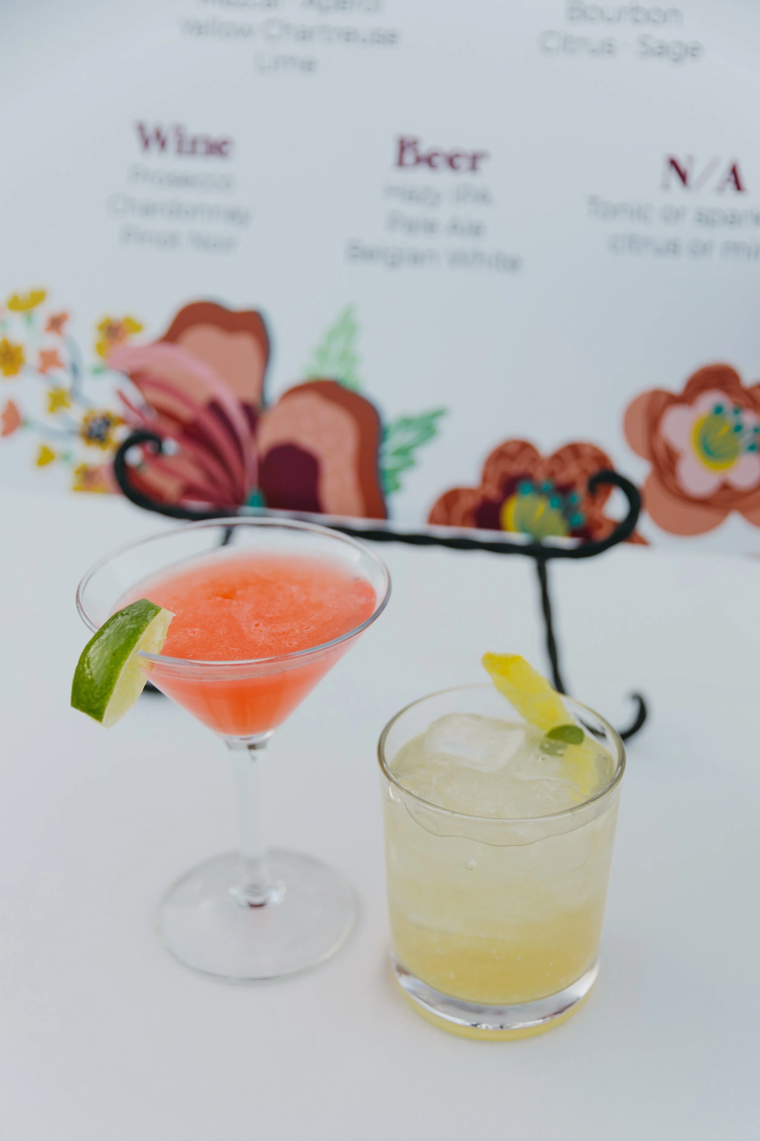 Two cocktails, one pink with lime wedge on a martini glass, and one yellow with lemon slice on a short glass, placed on a white surface.