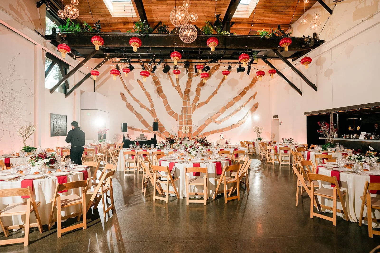 Wedding or banquet reception setup in a decorated hall featuring round tables with white tablecloths, red accents, floral centerpieces, wooden chairs, a large wall mural of a tree painted in brown, and hanging red lanterns with warm lighting.