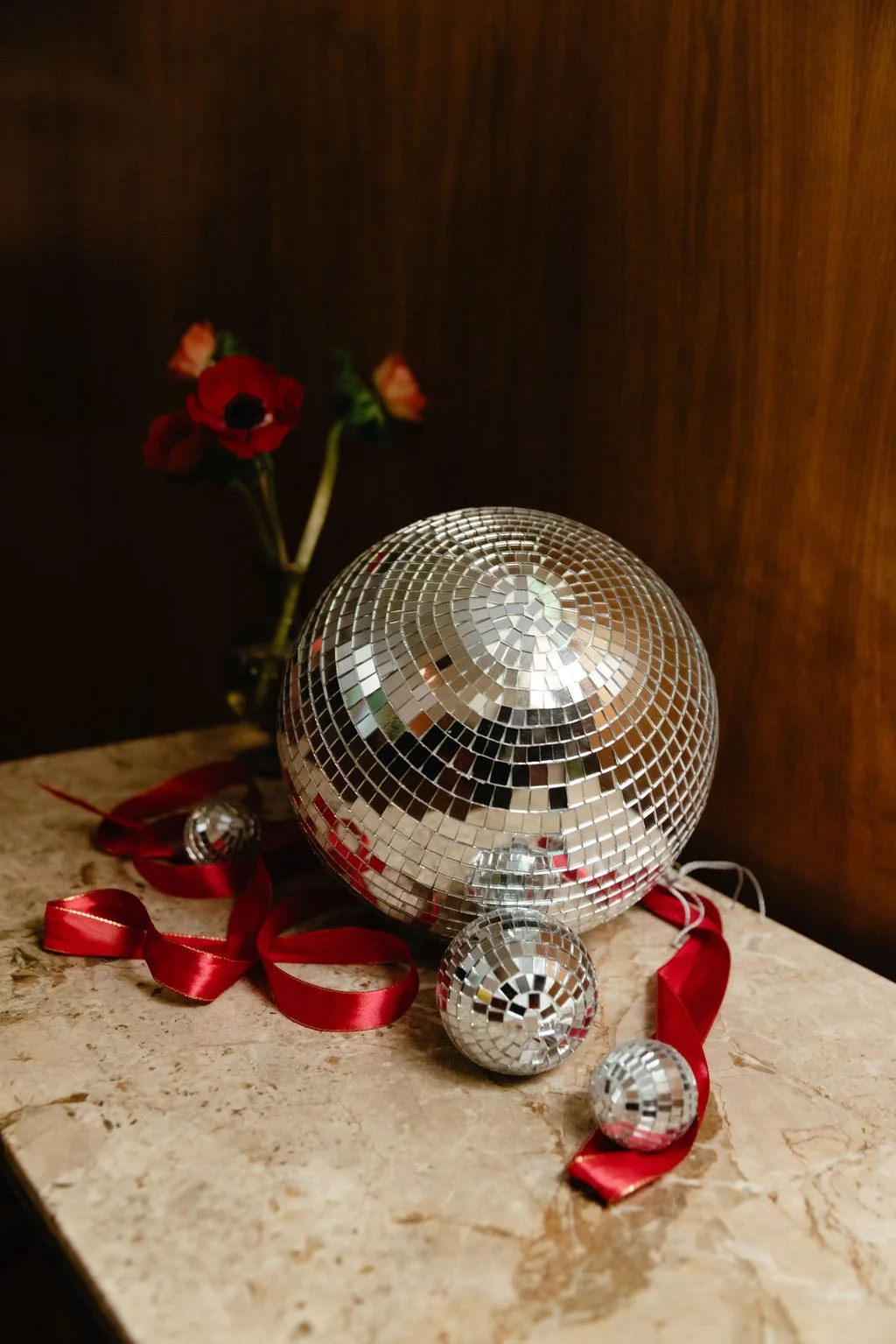 Disco ball and smaller disco balls on a table with red ribbon, with a vase of red flowers in the background against a wooden wall.