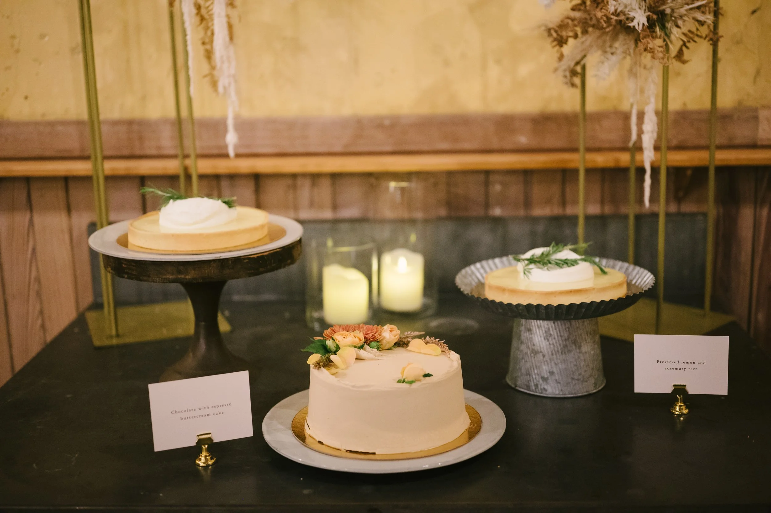 Three cakes displayed on a dark table with candles in the background. The center cake has flowers and is labeled 'Chocolate with espresso buttercream cake,' the other two cakes are on elevated stands, one with a lemon and rosemary tart, the other wit