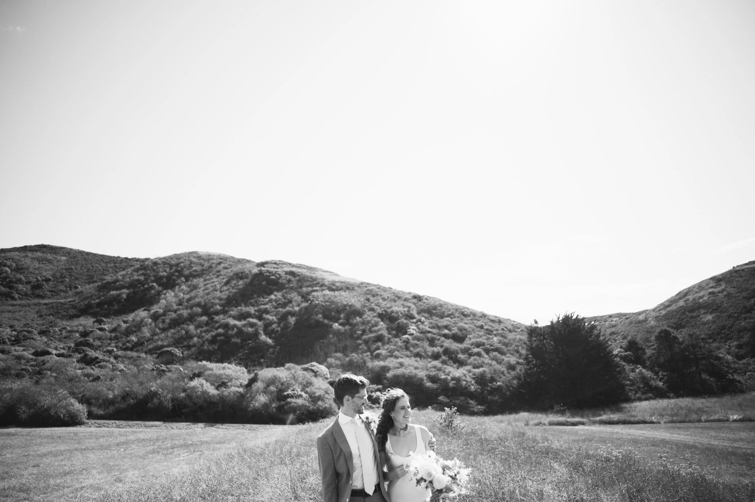 A black and white photo of a newlywed couple standing in a field with rolling hills in the background. The bride is holding a bouquet and the groom has his arm around her.