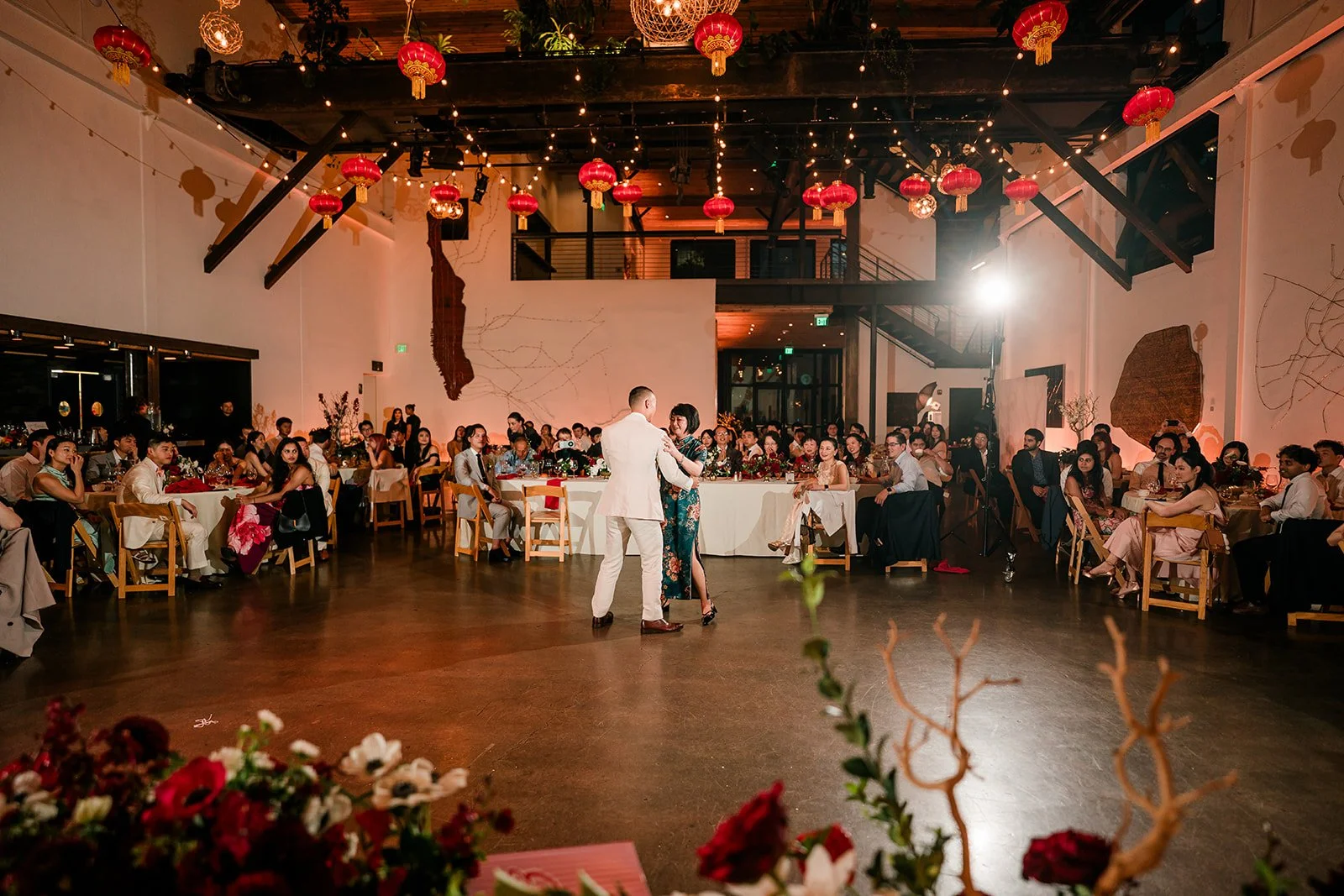 A couple is dancing in the center of a decorated banquet hall during a wedding reception, surrounded by seated guests at tables, with artwork and string lights on the walls and red lanterns hanging from the ceiling.