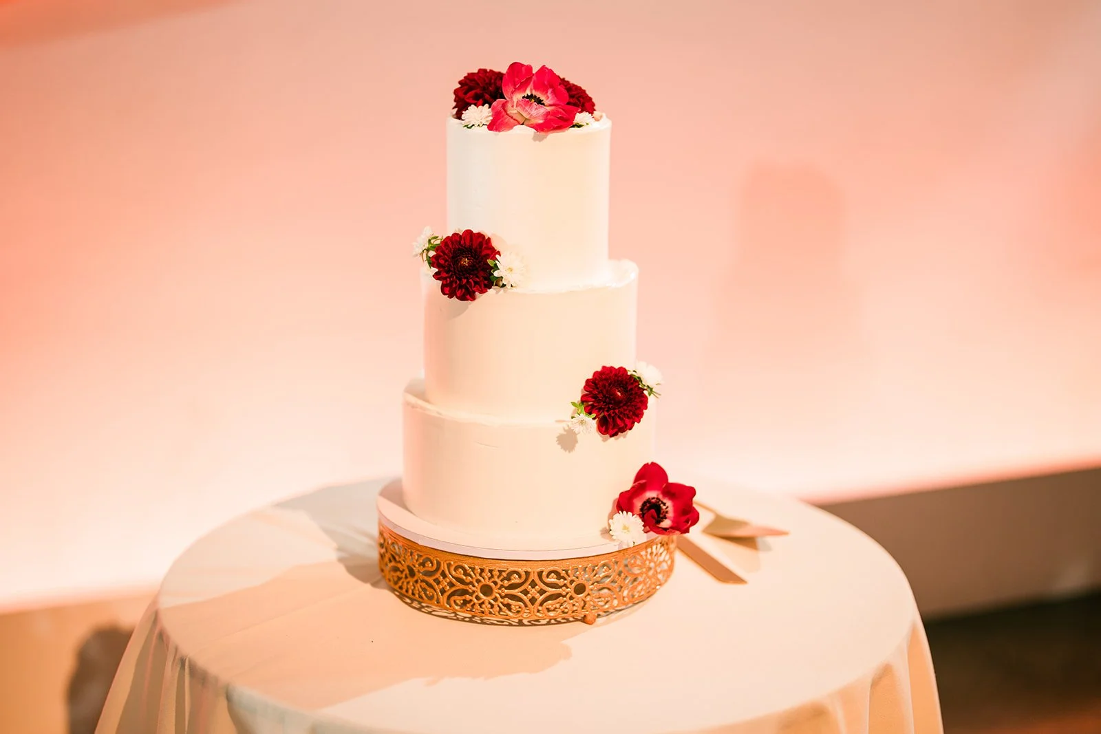 A three-tier white wedding cake decorated with red and white flowers, placed on a decorative gold cake stand on a round table with a cream-colored tablecloth.