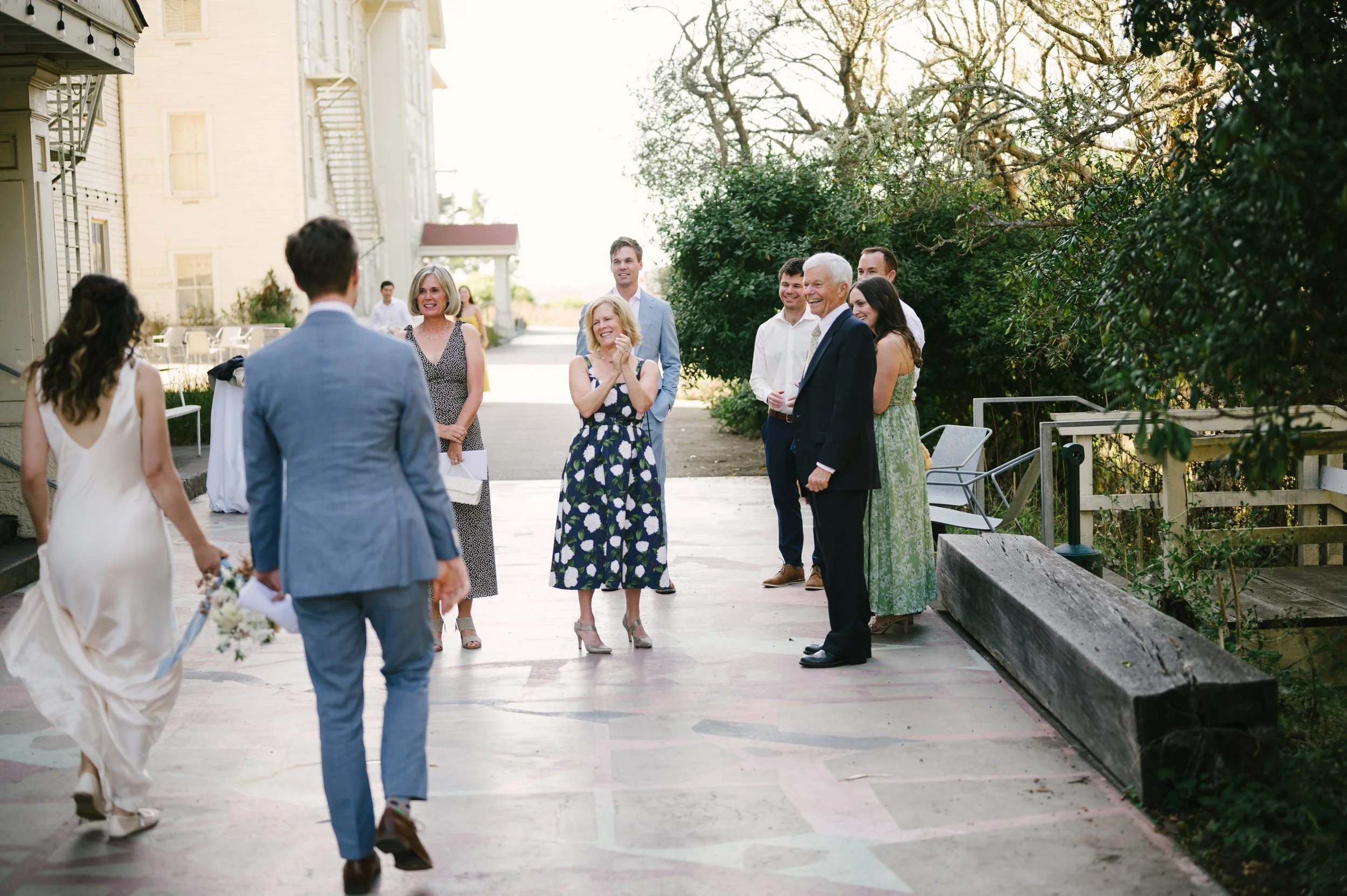 People gathered outdoors at The Marin Headlands Center for the Arts, celebrating a wedding, with a woman in a white dress holding a bouquet and a man in a blue suit walking towards her.