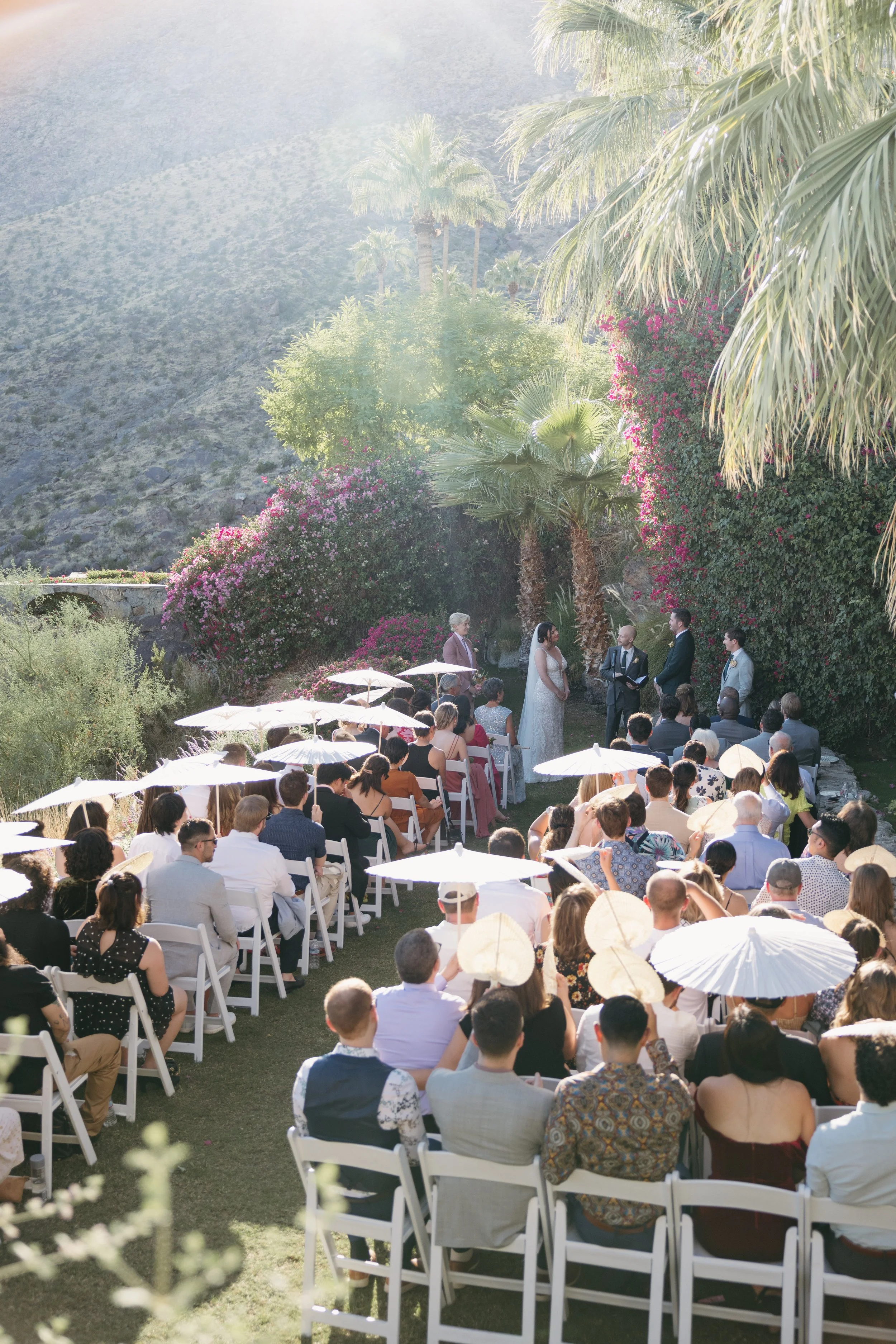 Outdoor wedding ceremony with guests seated on white chairs, holding umbrellas, facing an officiant and a bride and groom, set against a backdrop of blooming flowers and palm trees in a sunny, mountainous area.