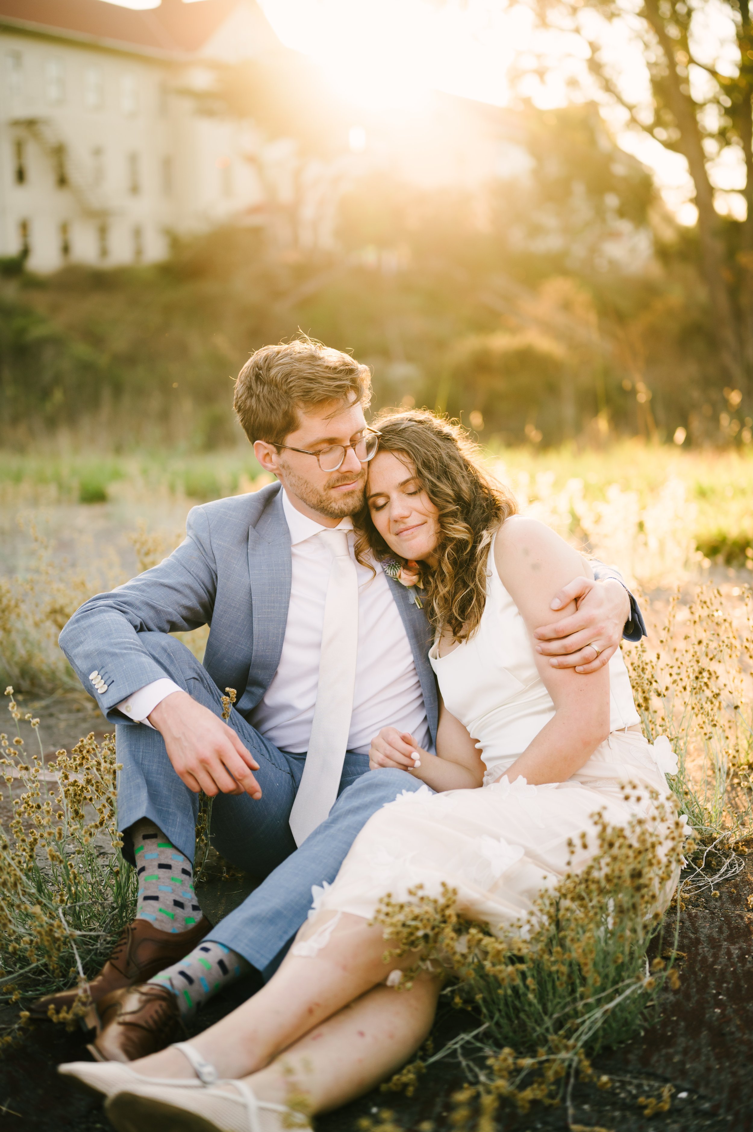 A couple sitting together outdoors in a field during sunset, embracing each other peacefully with their eyes closed.