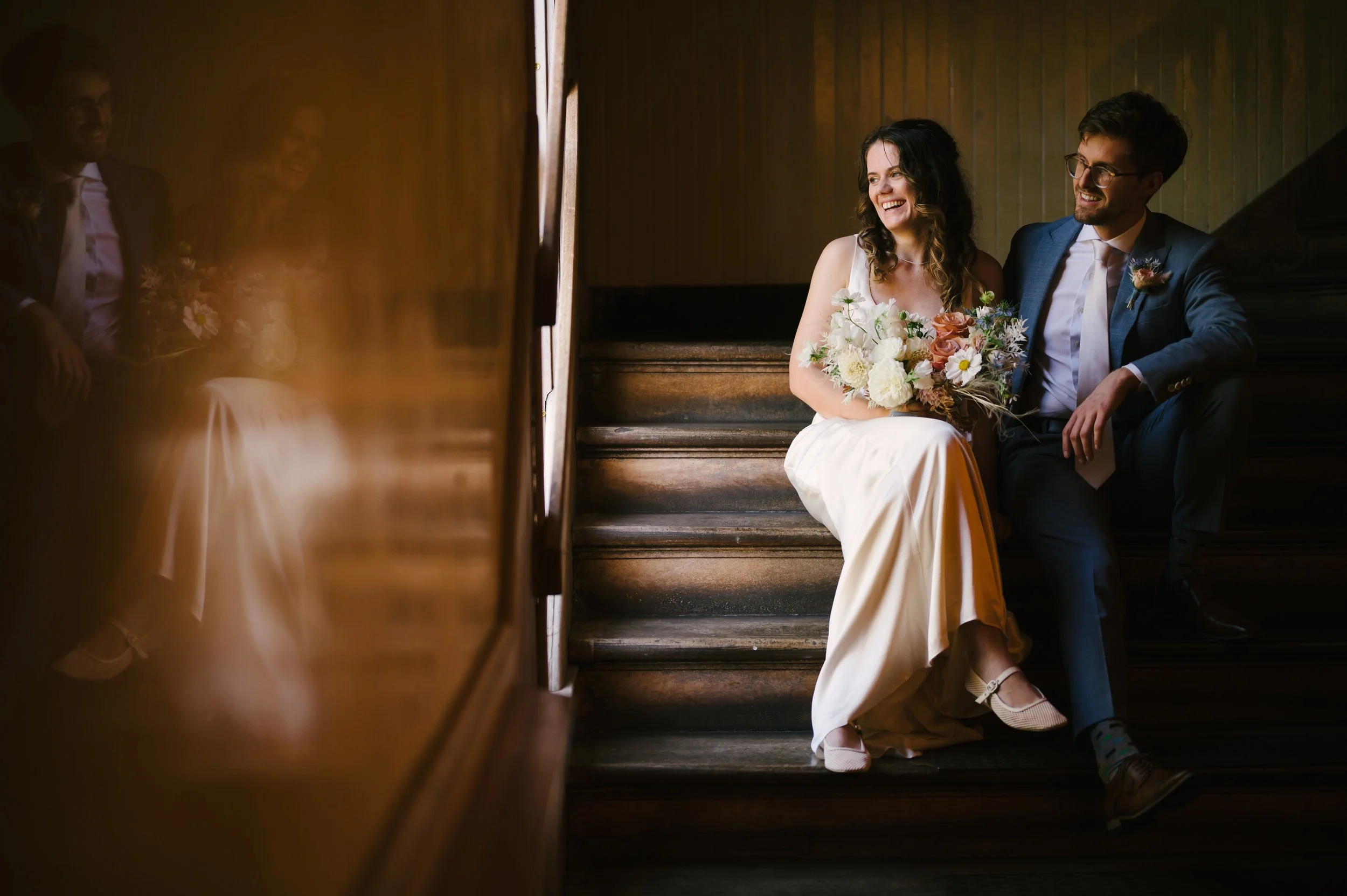 A bride and groom sitting on wooden stairs in the Marin Headlands Center for the Arts, smiling and looking at each other. The bride holds a bouquet of flowers and wears a white dress, while the groom is dressed in a blue suit. 