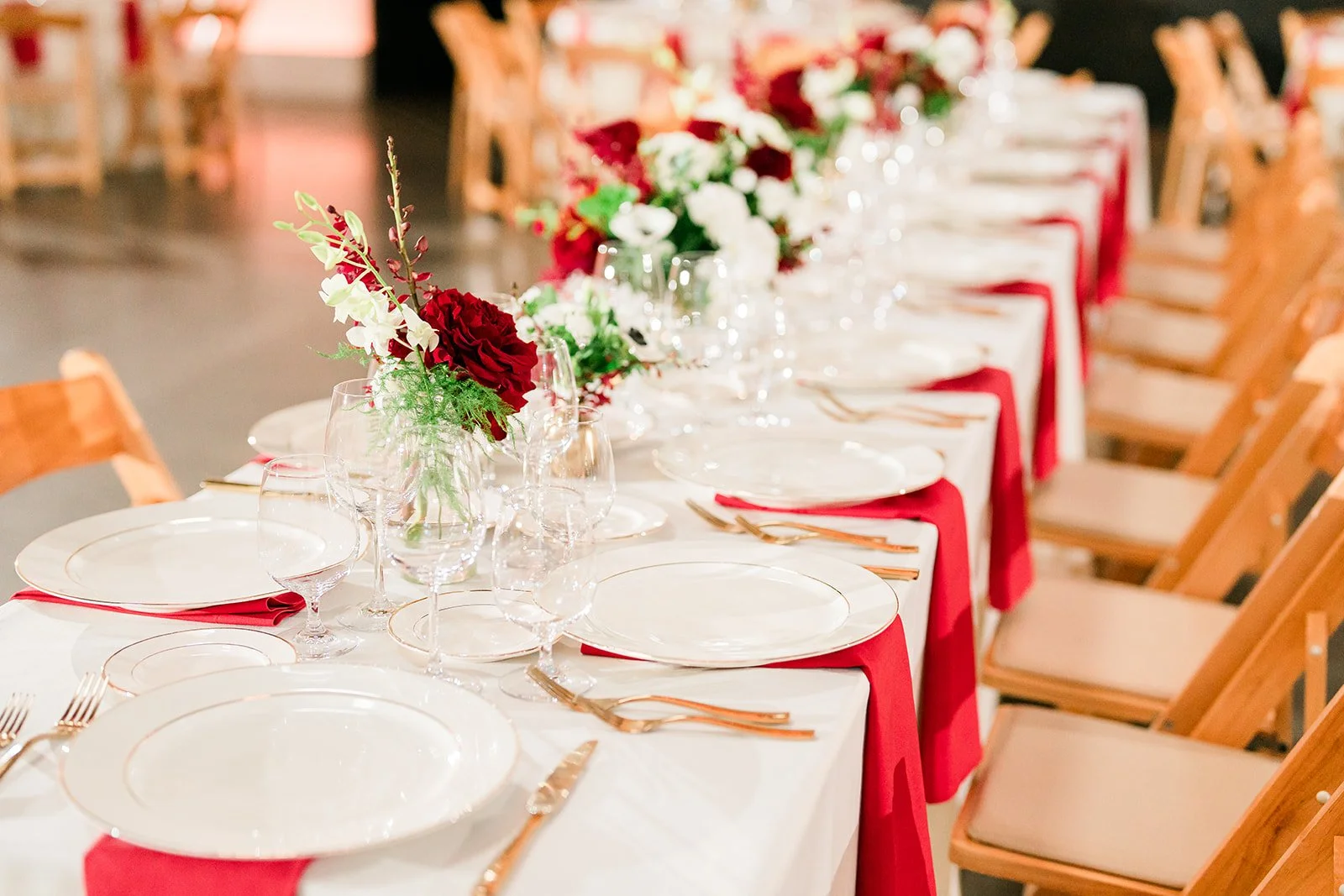 A long dining table set for a formal event with white tablecloths, red napkins, gold utensils, and glassware. There are floral centerpieces with red and white flowers along the table. Wooden chairs with beige cushions surround the table.