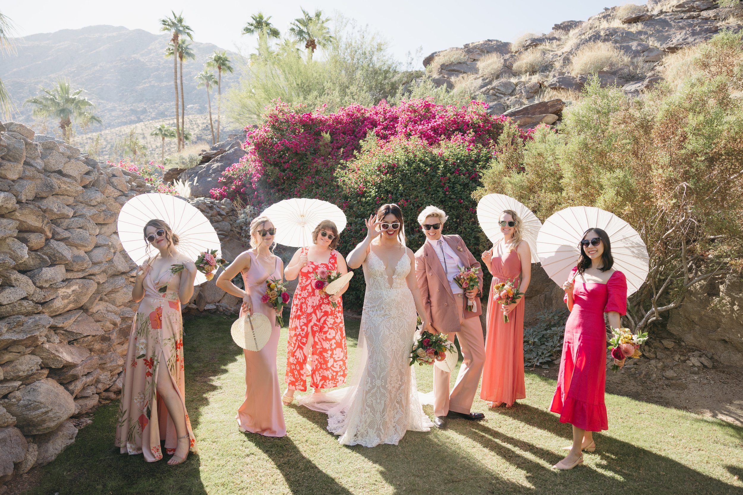 A group of women, including a bride in a white wedding dress, posing outdoors in a sunny garden with rocky terrain and pink flowering bushes, holding umbrellas and bouquets.