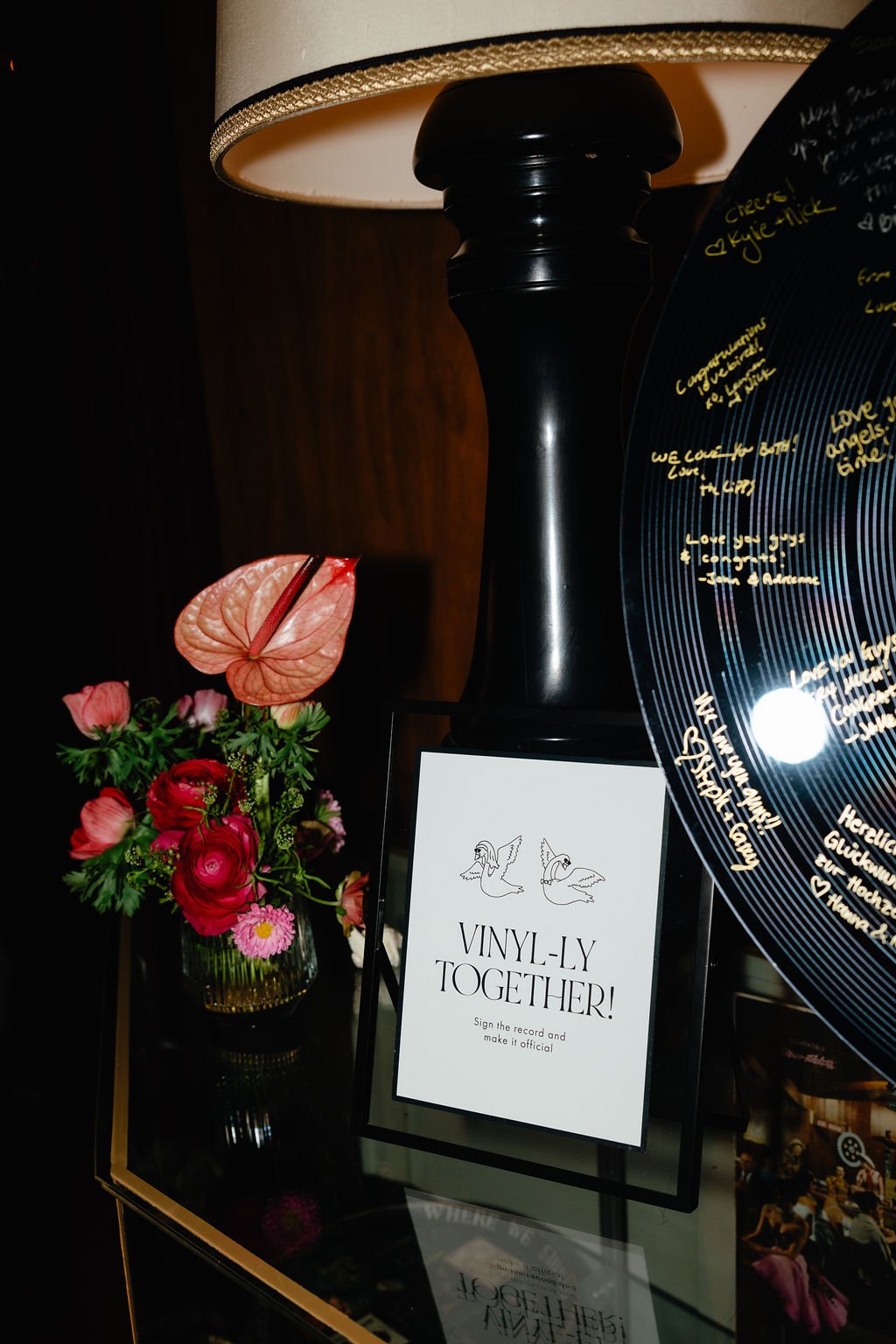 A floral arrangement with pink and red flowers next to a sign that reads 'VINYL-LY TOGETHER! Sign the record and make it official' with illustrations of two swans, set on a table with a black lamp in the background and handwritten messages on a vinyl