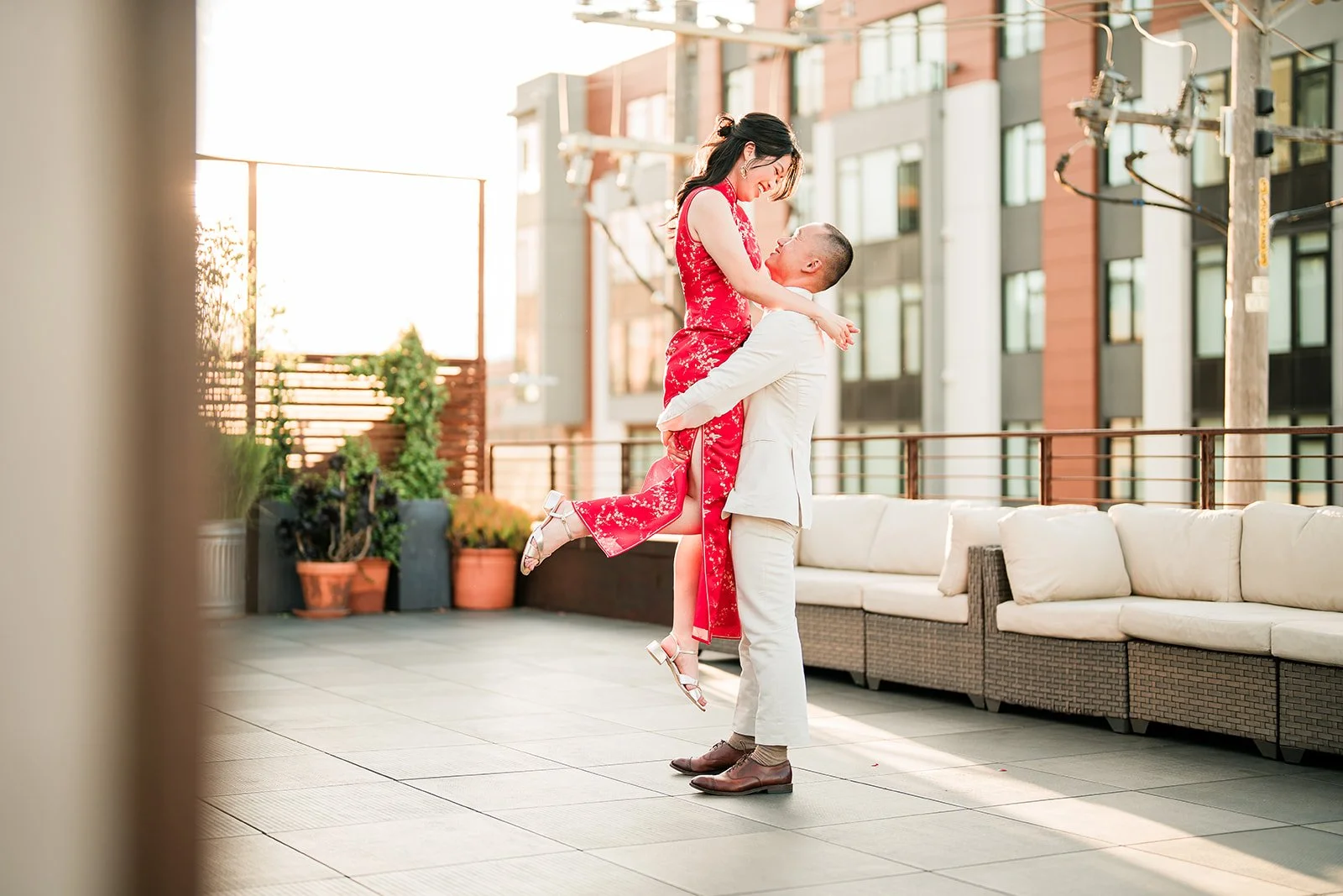 A man lifting a woman in a red dress with gold patterns on the rooftop terrace of The Pearl SF during sunset, with plants and modern apartment buildings in the background.