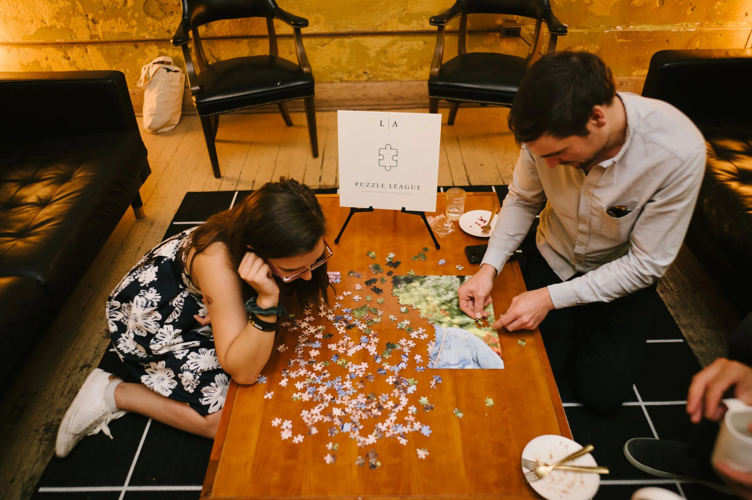 Two people working on assembling a jigsaw puzzle on a wooden table in a cozy room. The puzzle features a nature scene. There are puzzle pieces scattered on the table, a sign that says 'Puzzle League,' and some plates and a glass of water.