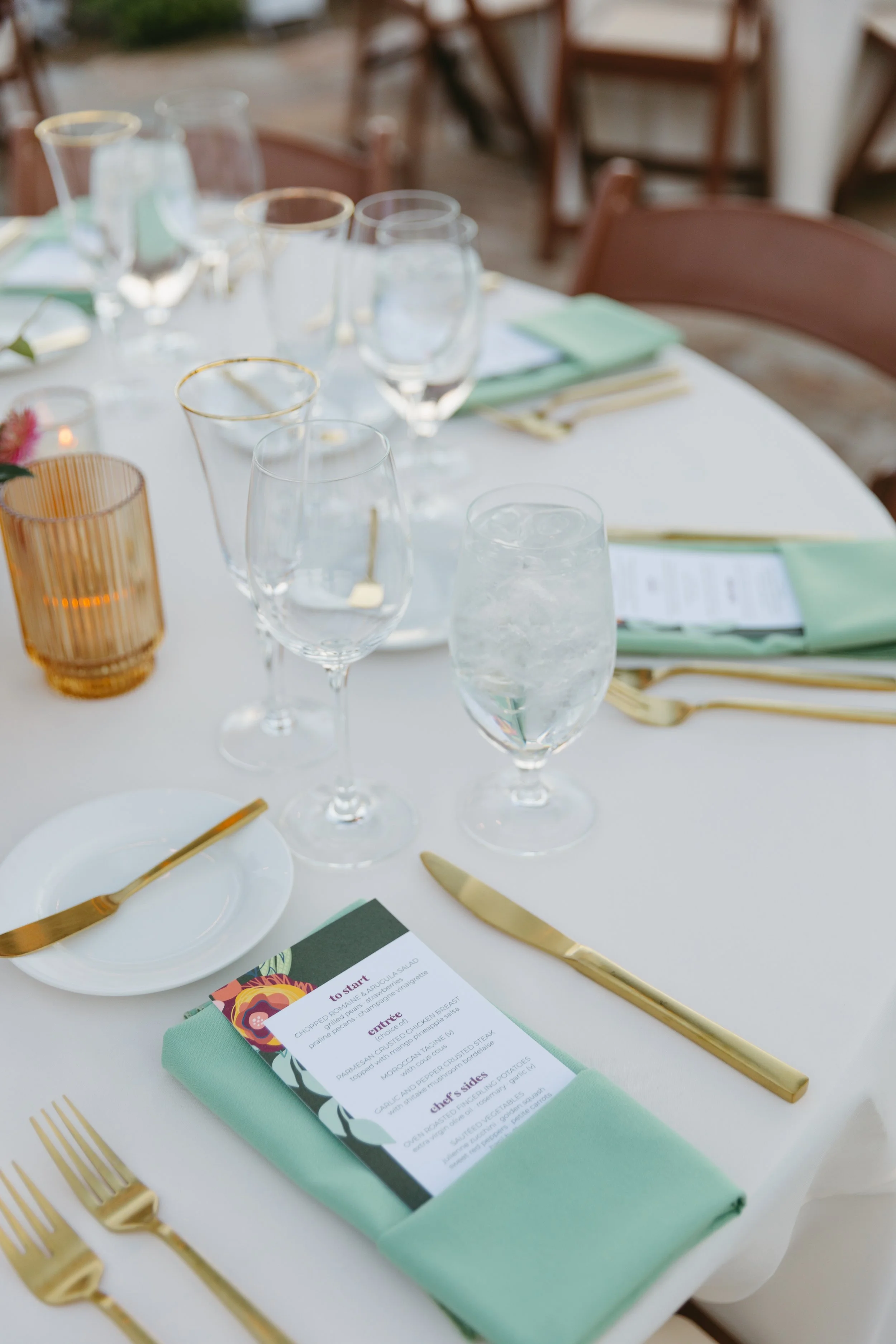Elegant dining table set with gold cutlery, green napkins, and a printed menu on a mint napkin, with glasses of water and wine, and candles on a white tablecloth.